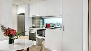 A modern kitchen with white cabinetry, a stainless steel refrigerator, a microwave, and an oven. There is a dining table with chairs and a vase of flowers.