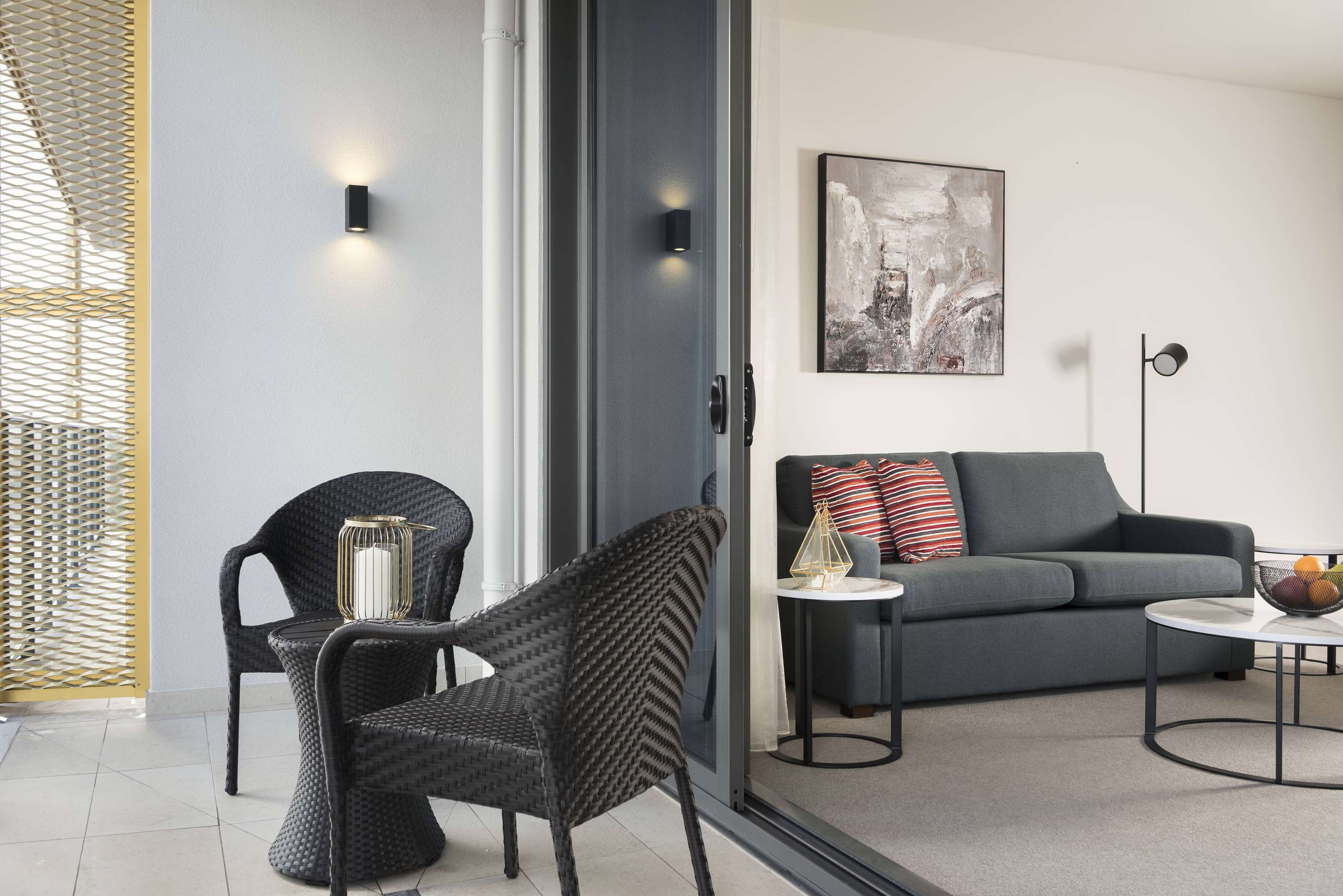 A modern living room with a grey sofa, two black chairs, a small table with a lamp, and a framed abstract painting on the wall.