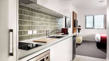 A modern kitchen with a built-in stove, a sink, and a countertop. There is a small dining area with a table and chairs. The room has a large window with sheer curtains.
