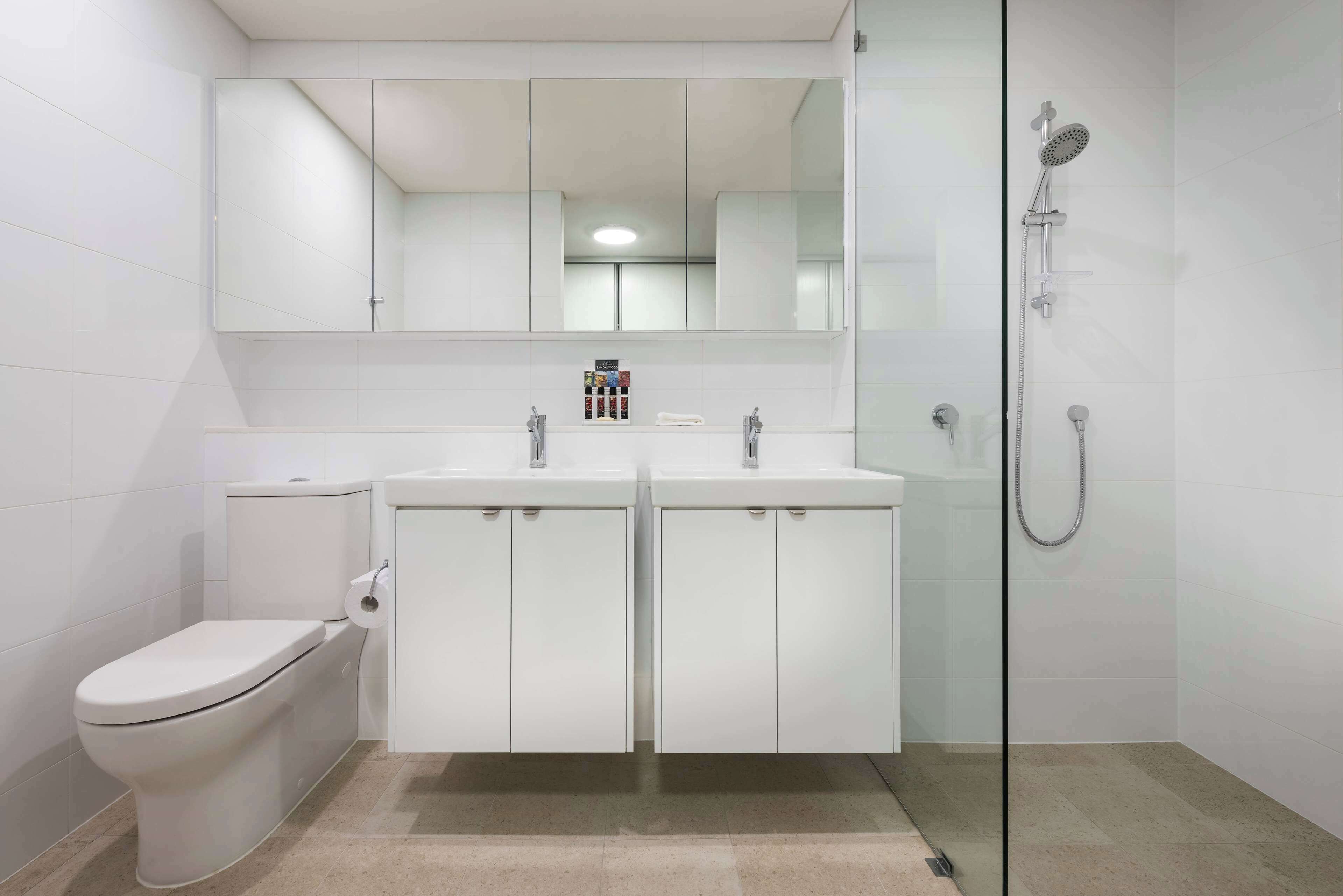 A modern bathroom with a toilet, two sinks, a shower, and a large mirror above the sinks.