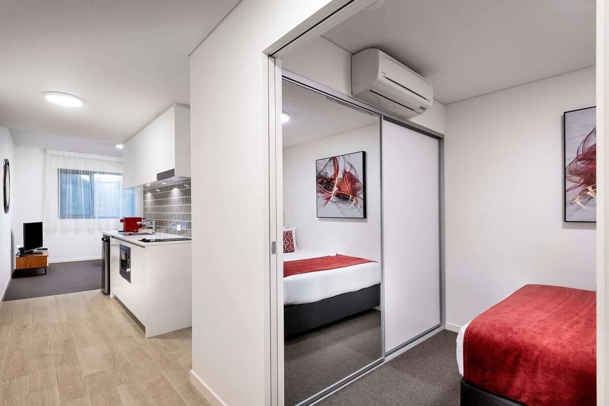A compact hotel room with a kitchenette, a bed with a red blanket, and a sliding door closet.