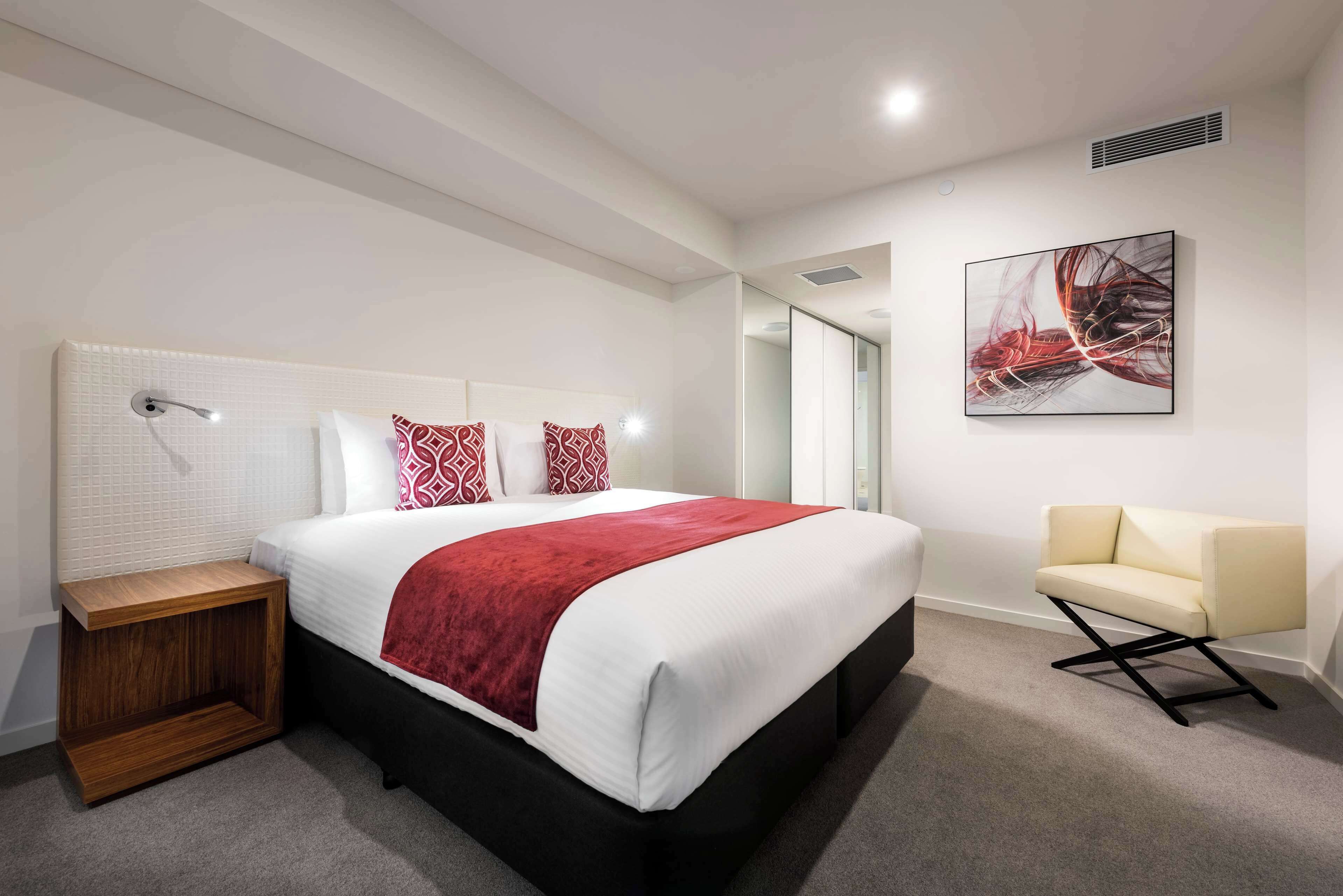 Une chambre d’hôtel moderne avec un grand lit, une table de chevet, une chaise et un tableau abstrait accroché au mur.