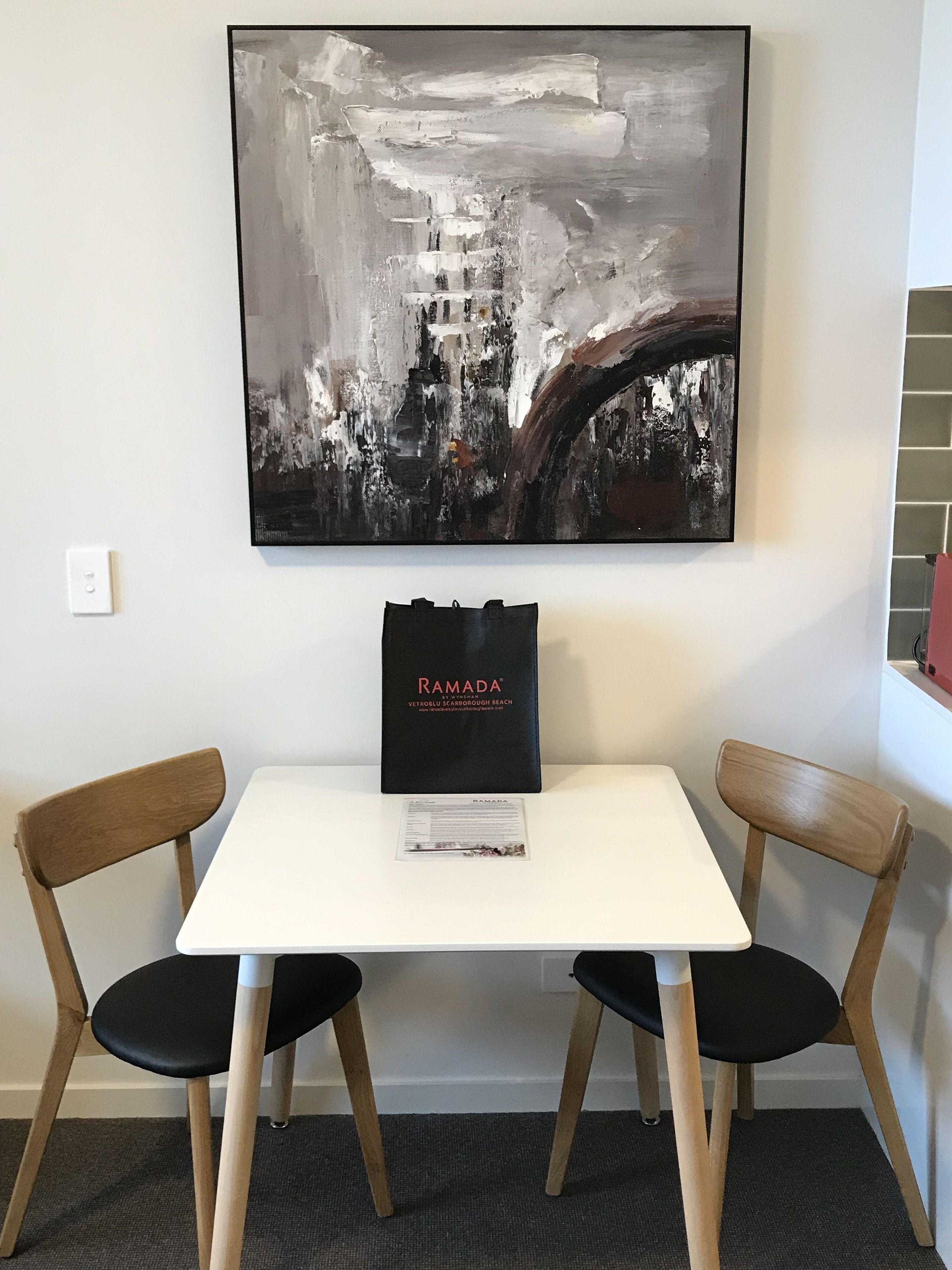 A white table with two wooden chairs, a black Ramada® tote bag, and a large abstract painting on the wall.