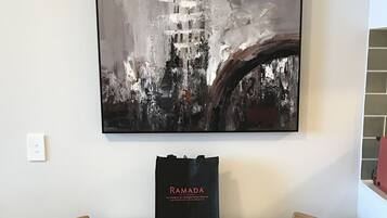 A white table with two wooden chairs, a black Ramada® tote bag, and a large abstract painting on the wall.