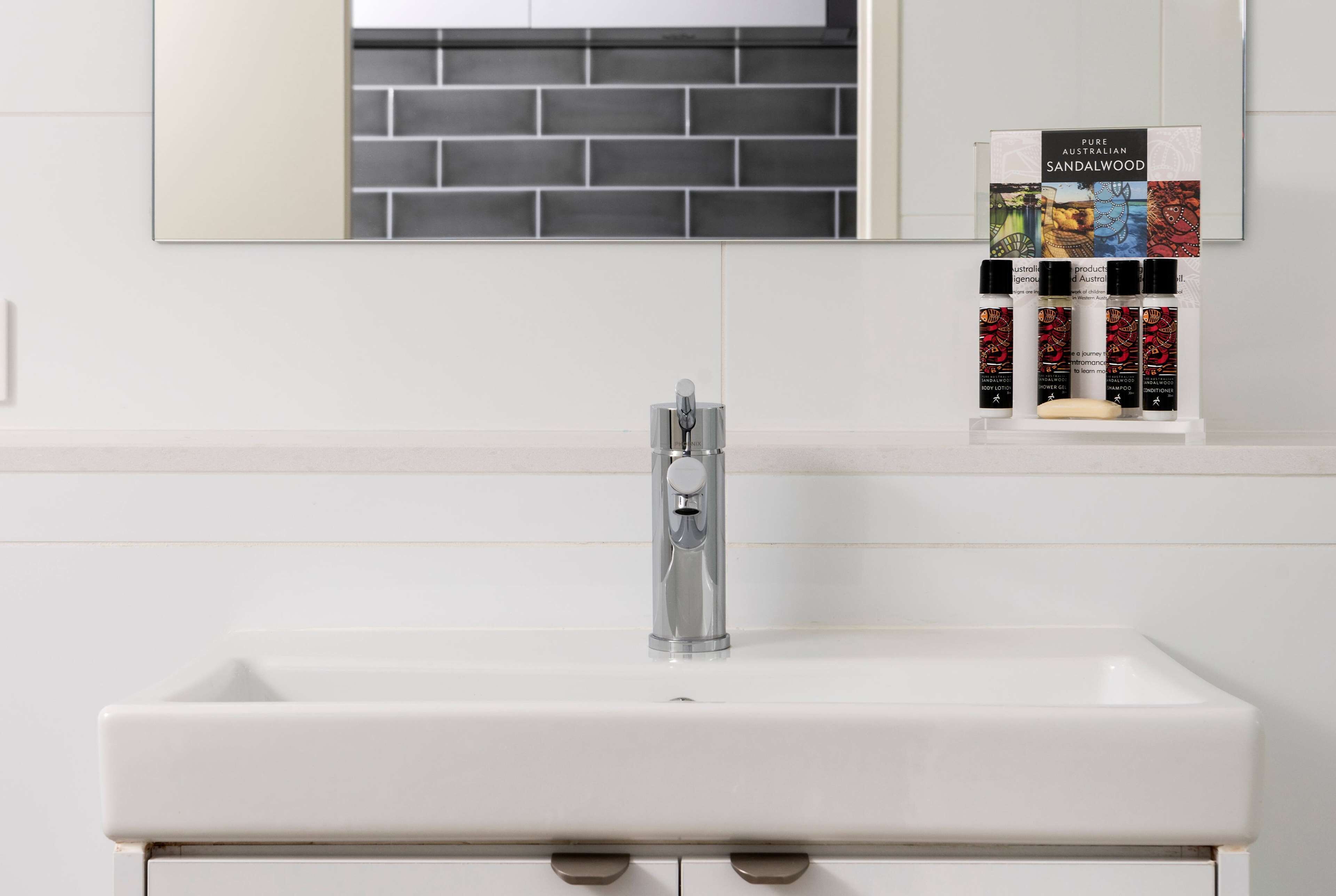 A bathroom with a white sink, a modern faucet, and a mirror above it. There is a shelf with a box labeled "Pure Australian Sandalwood" and several bottles of essential oils.