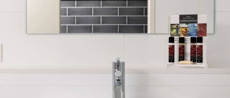 A bathroom with a white sink, a modern faucet, and a mirror above it. There is a shelf with a box labeled "Pure Australian Sandalwood" and several bottles of essential oils.