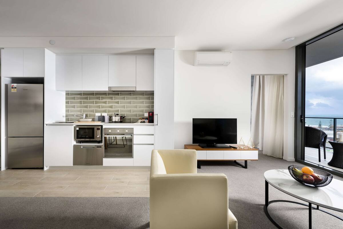A modern kitchen with white cabinets, a microwave, and a stove. There is a dining area with a table and chairs, and a TV on a stand. The room has a large window with a view of the sky.