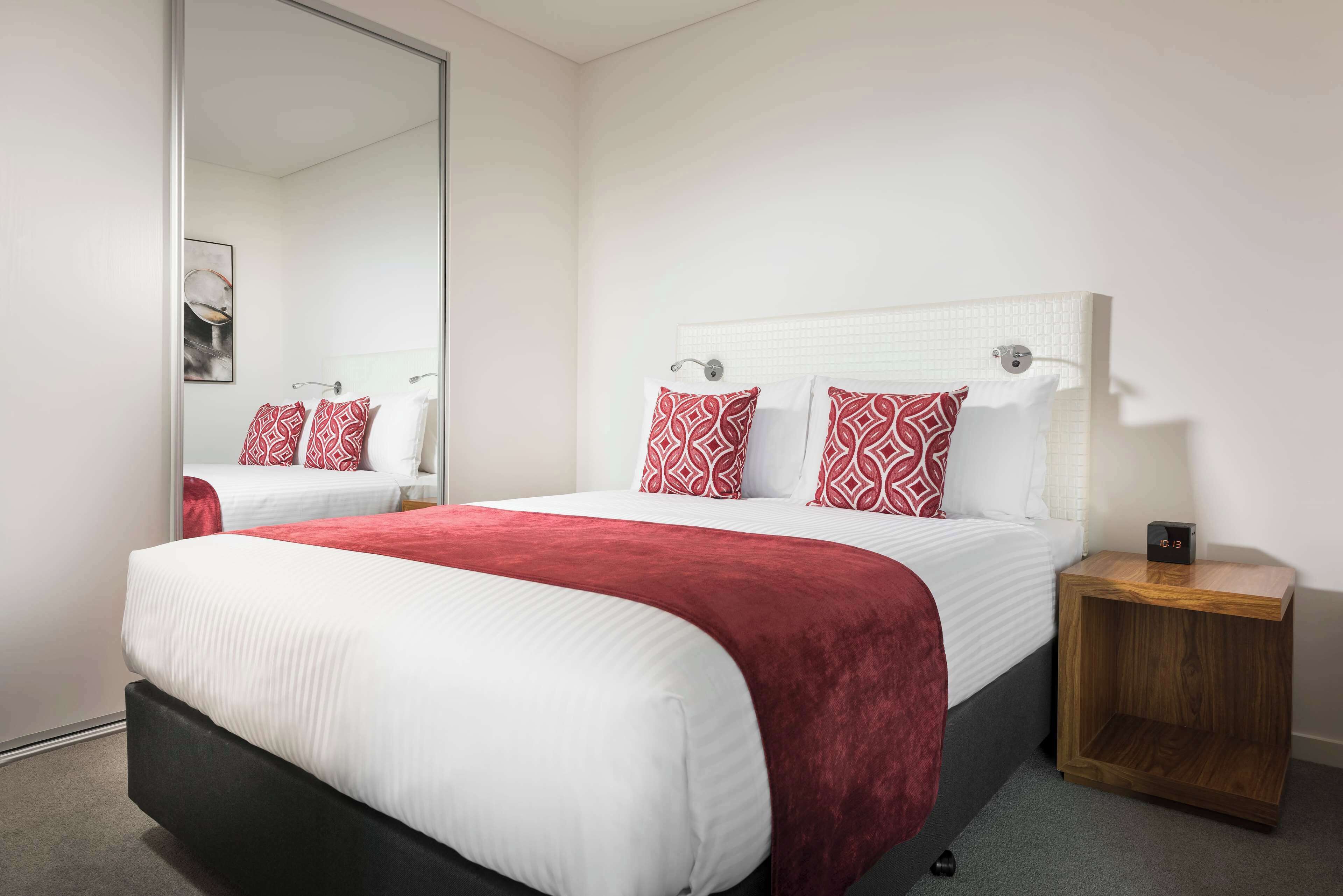 Une chambre d’hôtel avec un grand lit, une table de chevet en bois, un miroir et un coussin aux motifs rouges et blancs.