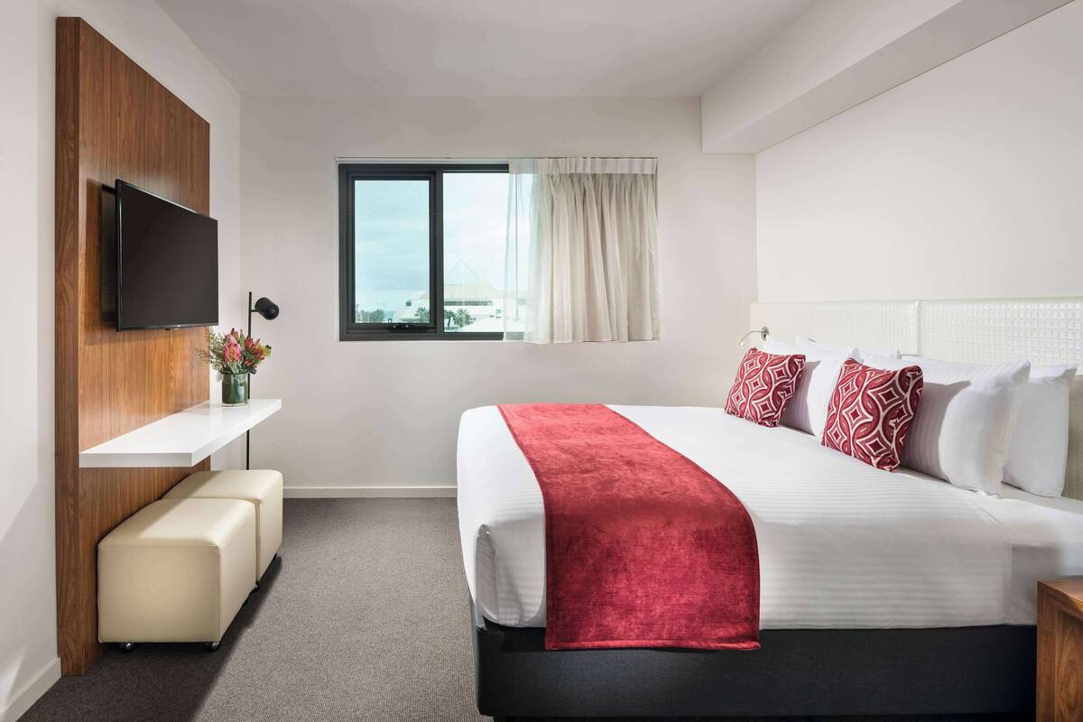 A hotel room with a bed, a TV mounted on a wooden panel, a window with curtains, a bench, and a nightstand.