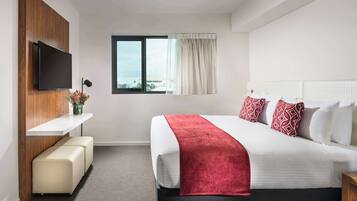 Habitación de hotel con cama, televisión montada en un panel de madera, ventana con cortinas, un banco y una mesita de noche.