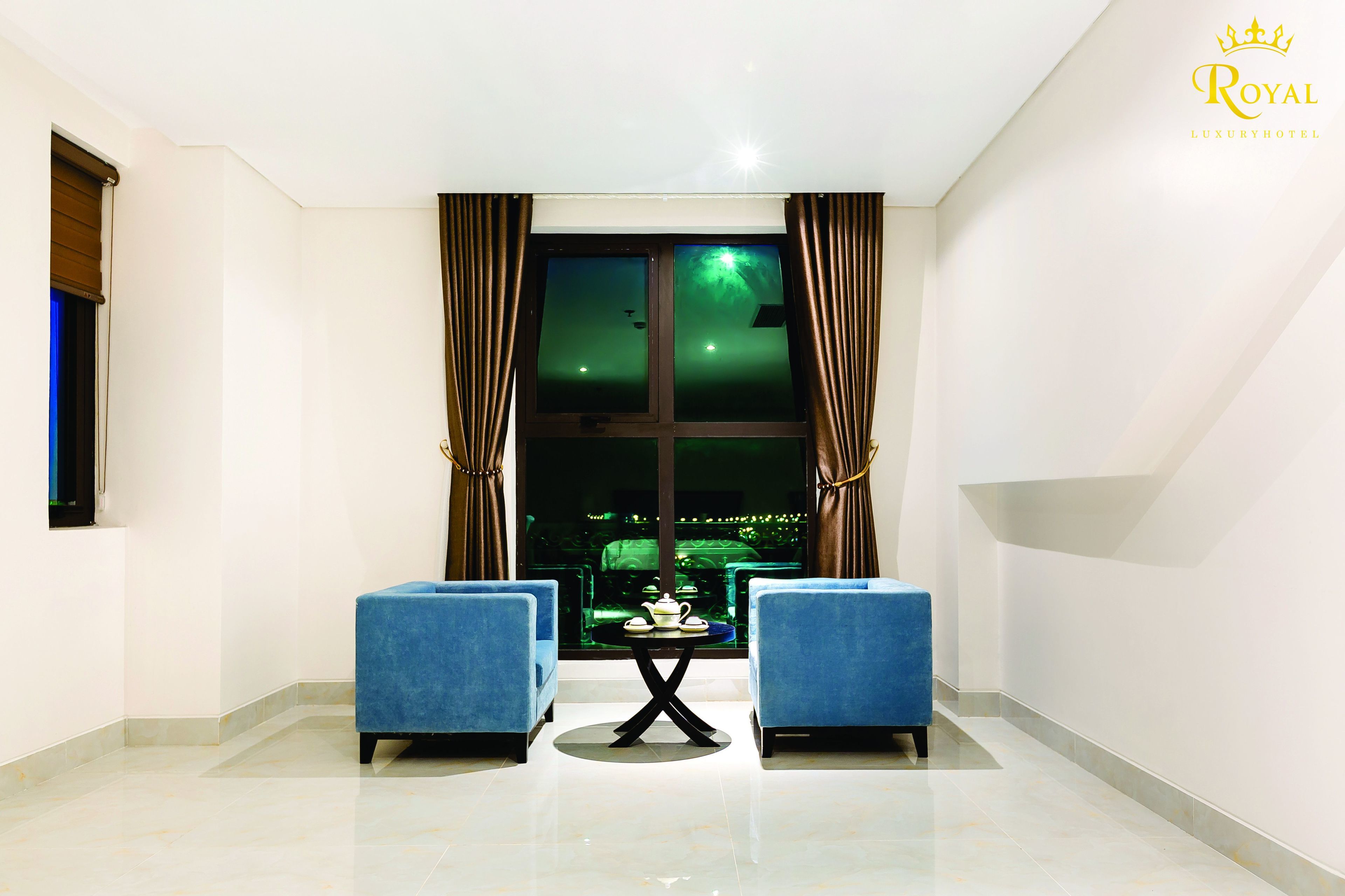 A modern room with two blue armchairs facing a table with a teapot and cups, a large window with curtains, and a view of a cityscape at night.