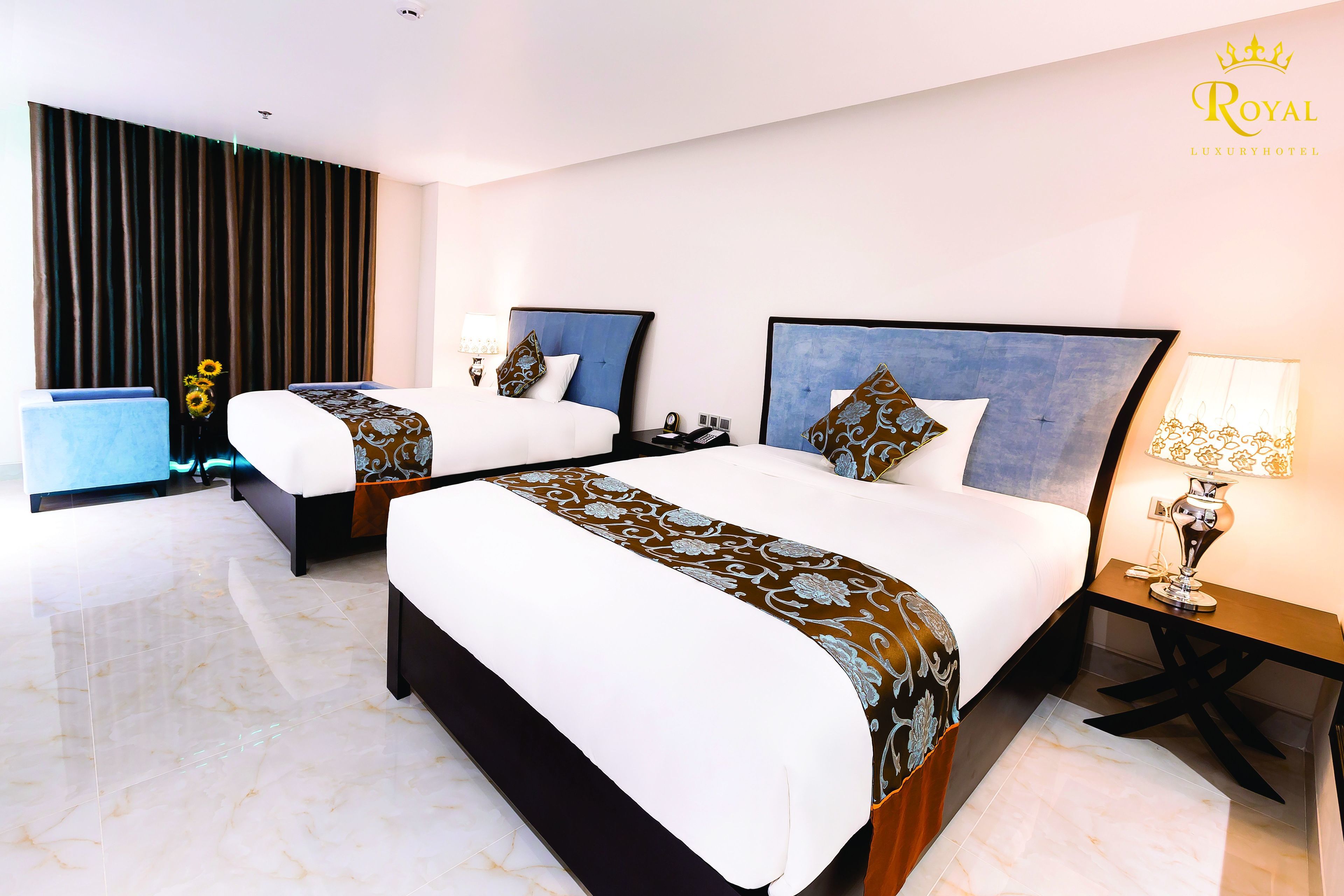 A hotel room with two beds, a large window with curtains, a nightstand with a lamp, and a vase with flowers.