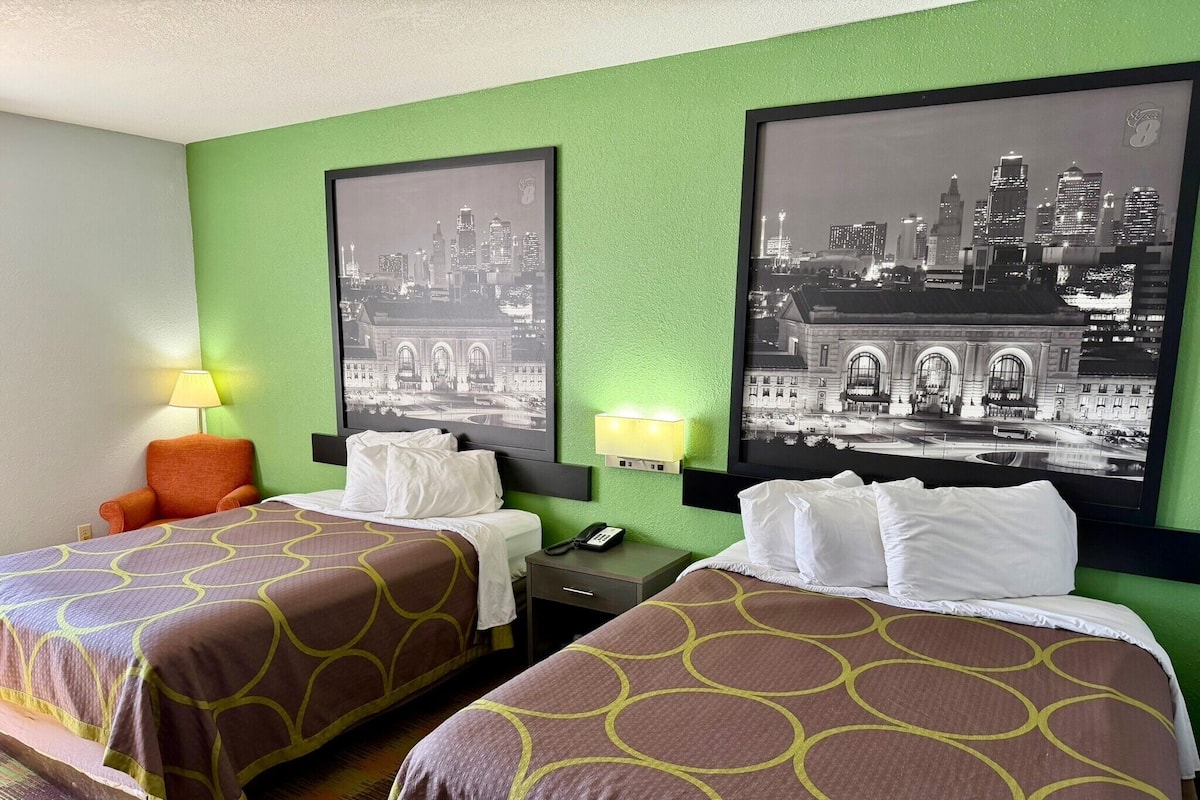 Habitación de hotel con dos camas, una mesita de noche, una lámpara y dos fotografías en blanco y negro del paisaje urbano enmarcadas en la pared.