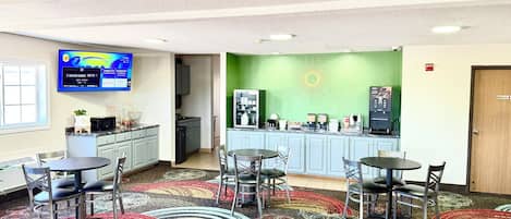 A hotel lobby with a kitchen area, a TV displaying sports channels, and a green wall with a sun design.