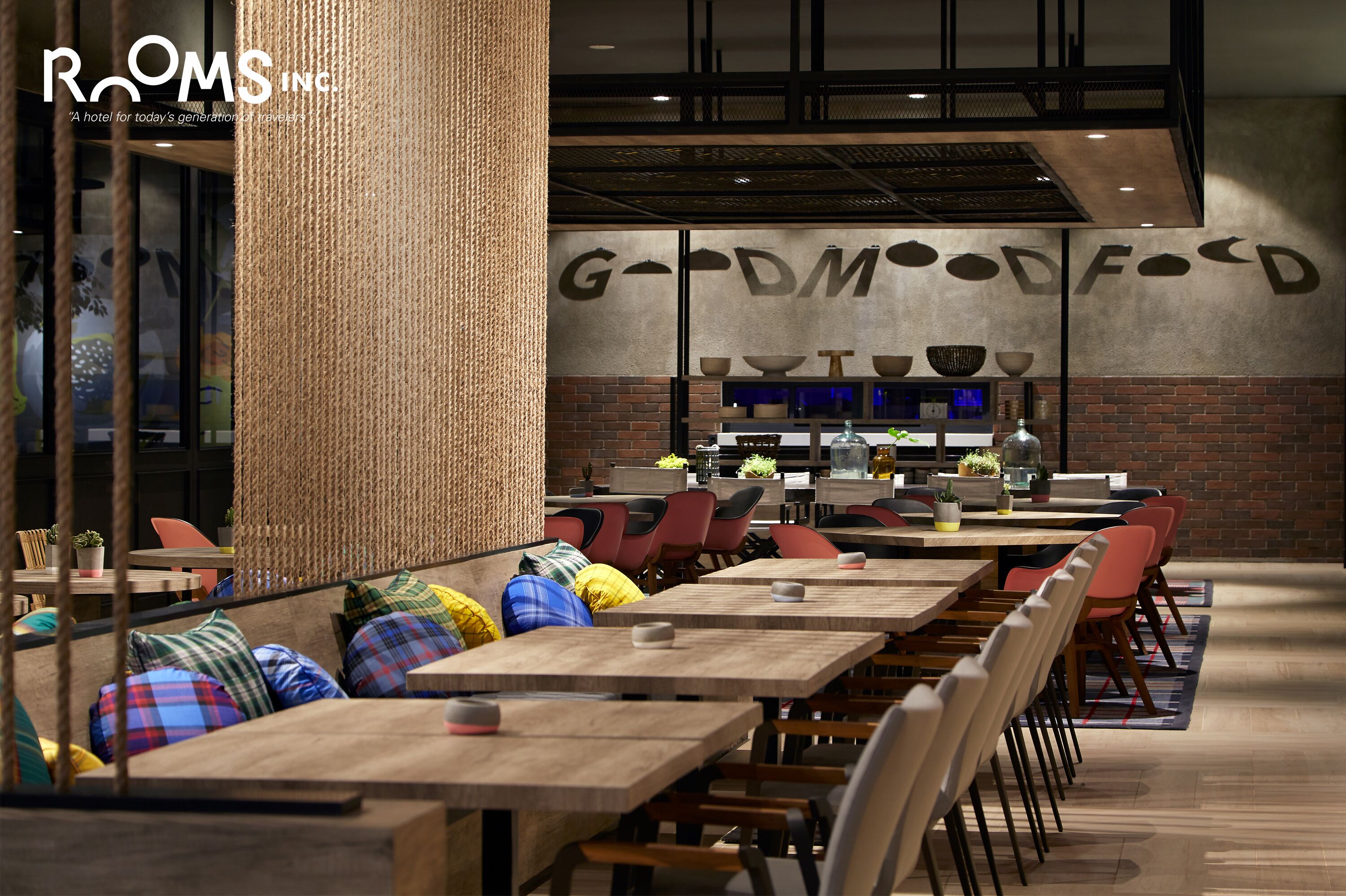 A modern restaurant with wooden tables and colorful cushions, a brick wall, and a 'G-DMOOOODE' sign.
