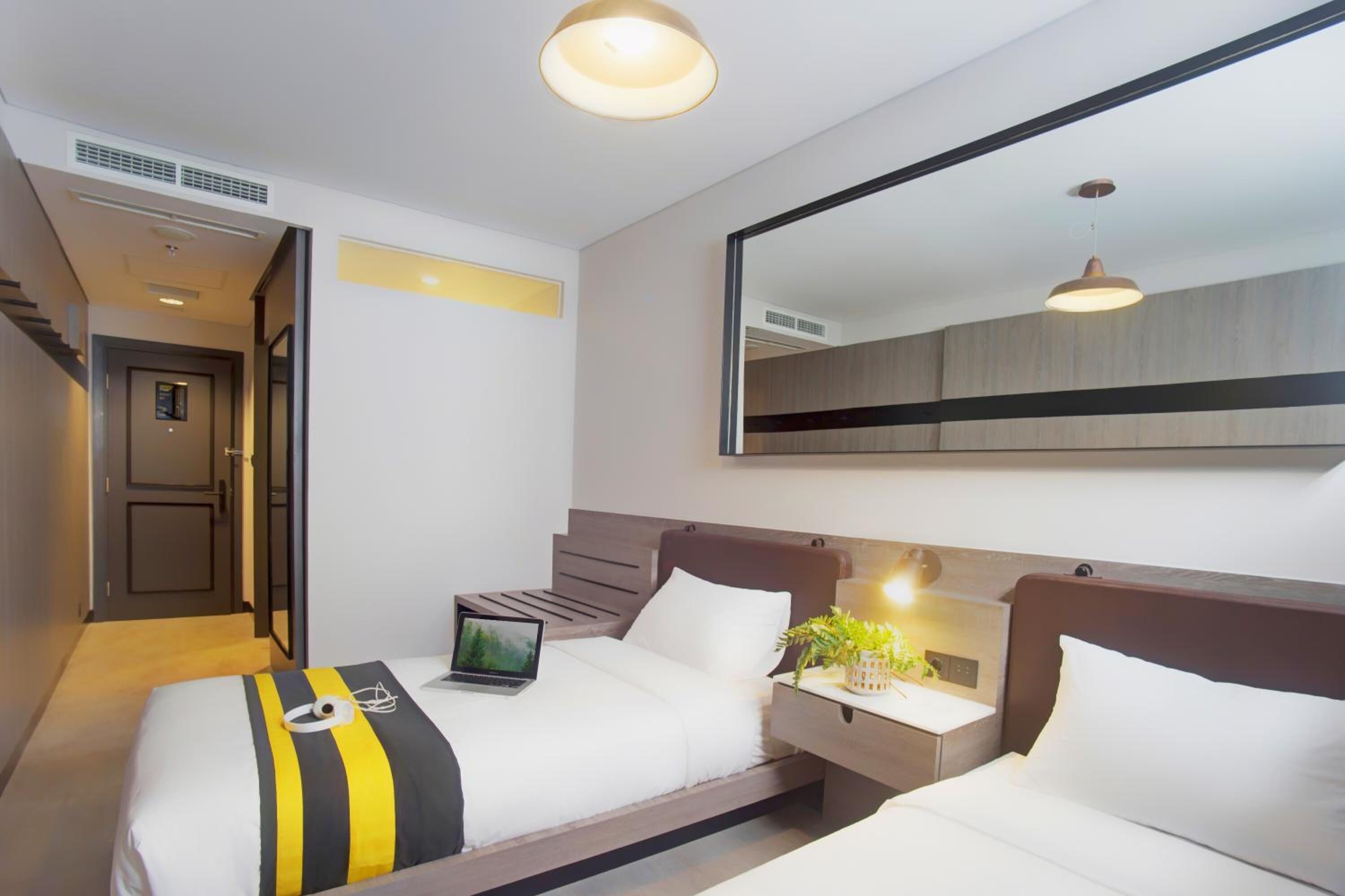 A hotel room with two beds, a large mirror, a bedside table with a lamp, and a laptop on one bed.