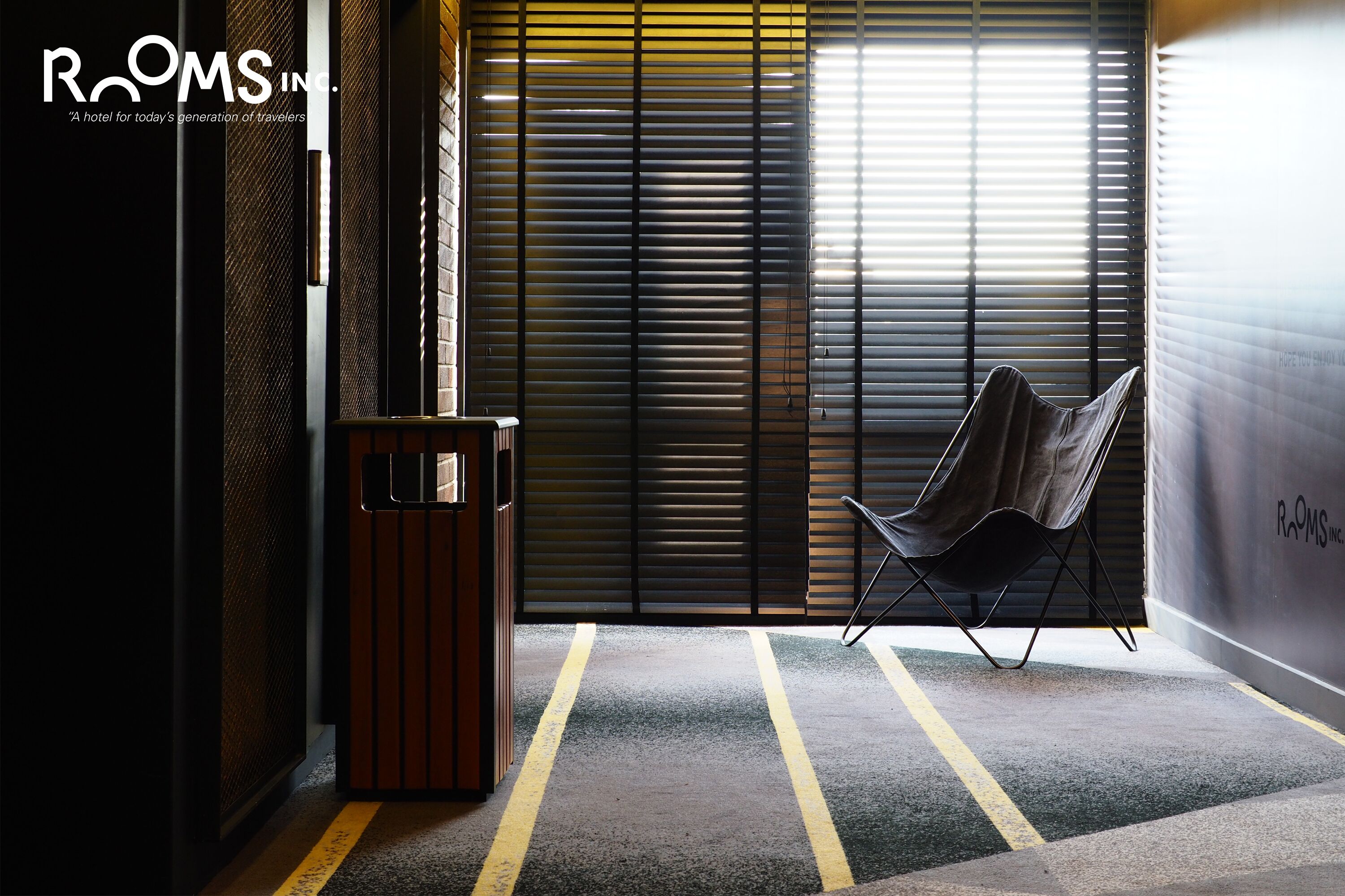 Un hall d’hôtel moderne avec une chaise à armature métallique, une poubelle et une grande fenêtre équipée de stores.