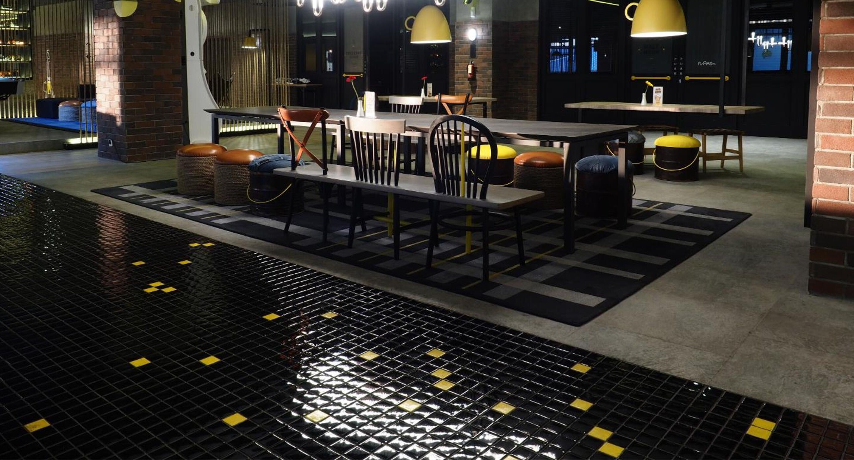 Un área de piscina interior moderna con suelo de baldosas negras y amarillas, rodeada de mesas y sillas con iluminación colgante amarilla.