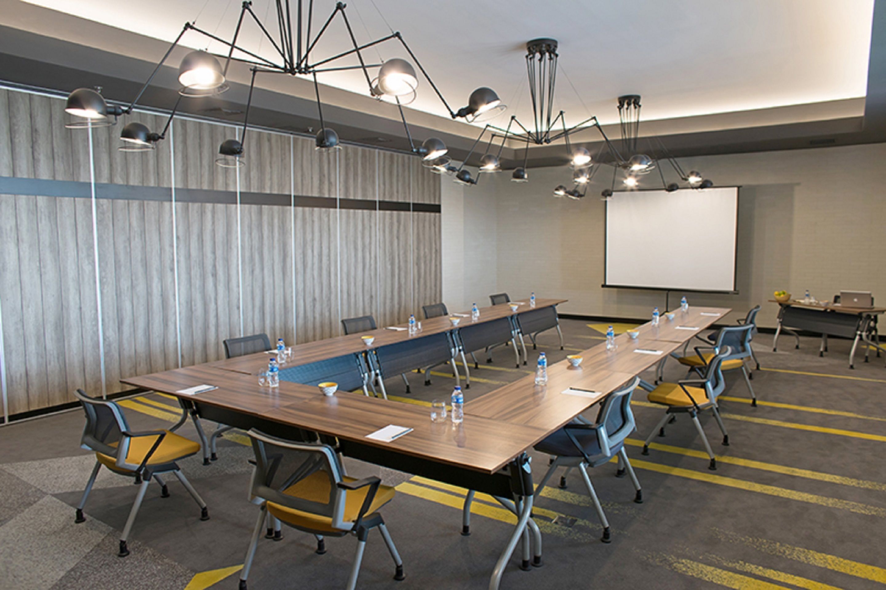 A conference room with a long table, chairs, a projector screen, and multiple ceiling lights.