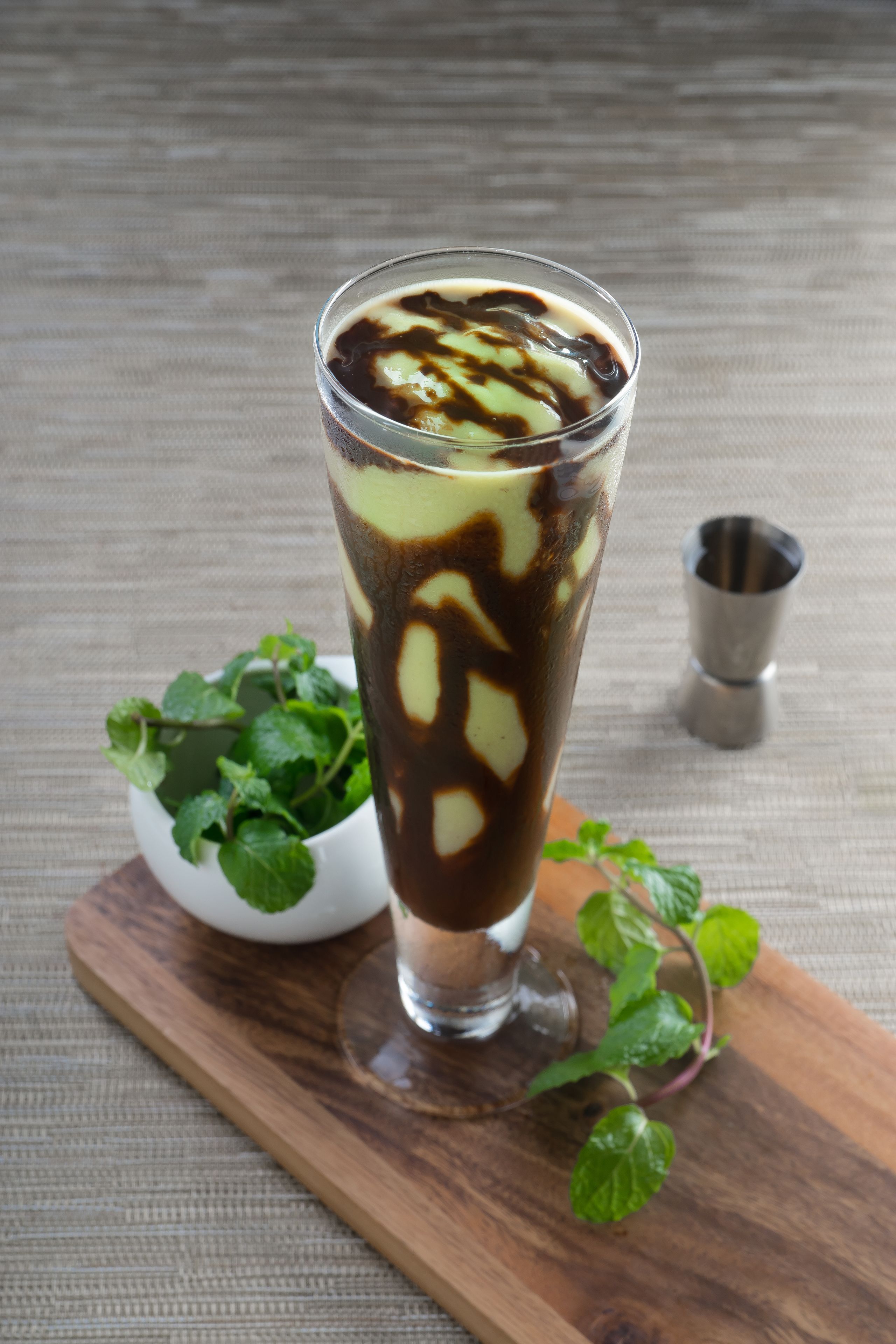 A tall glass of iced coffee with chocolate syrup, placed on a wooden board with mint leaves beside it.