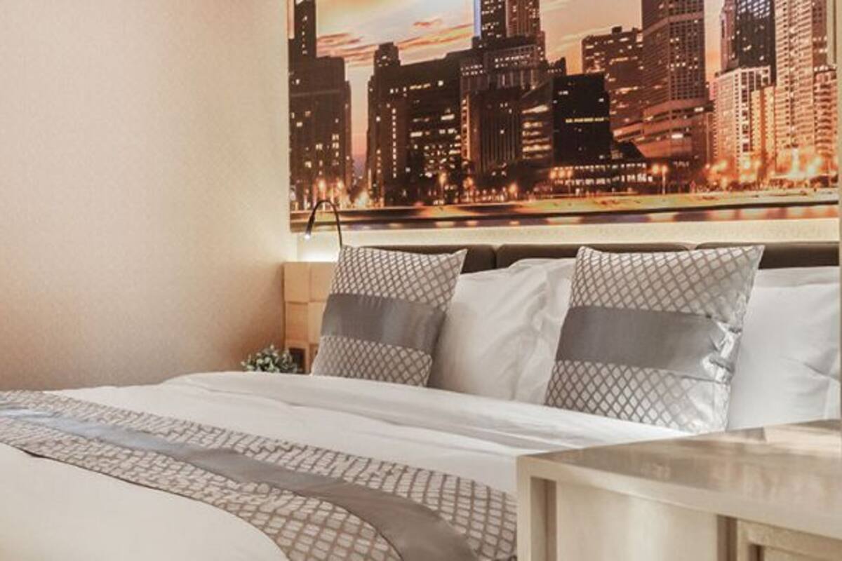 A hotel room with a bed, a nightstand with a vase of flowers, and a cityscape view on the wall.