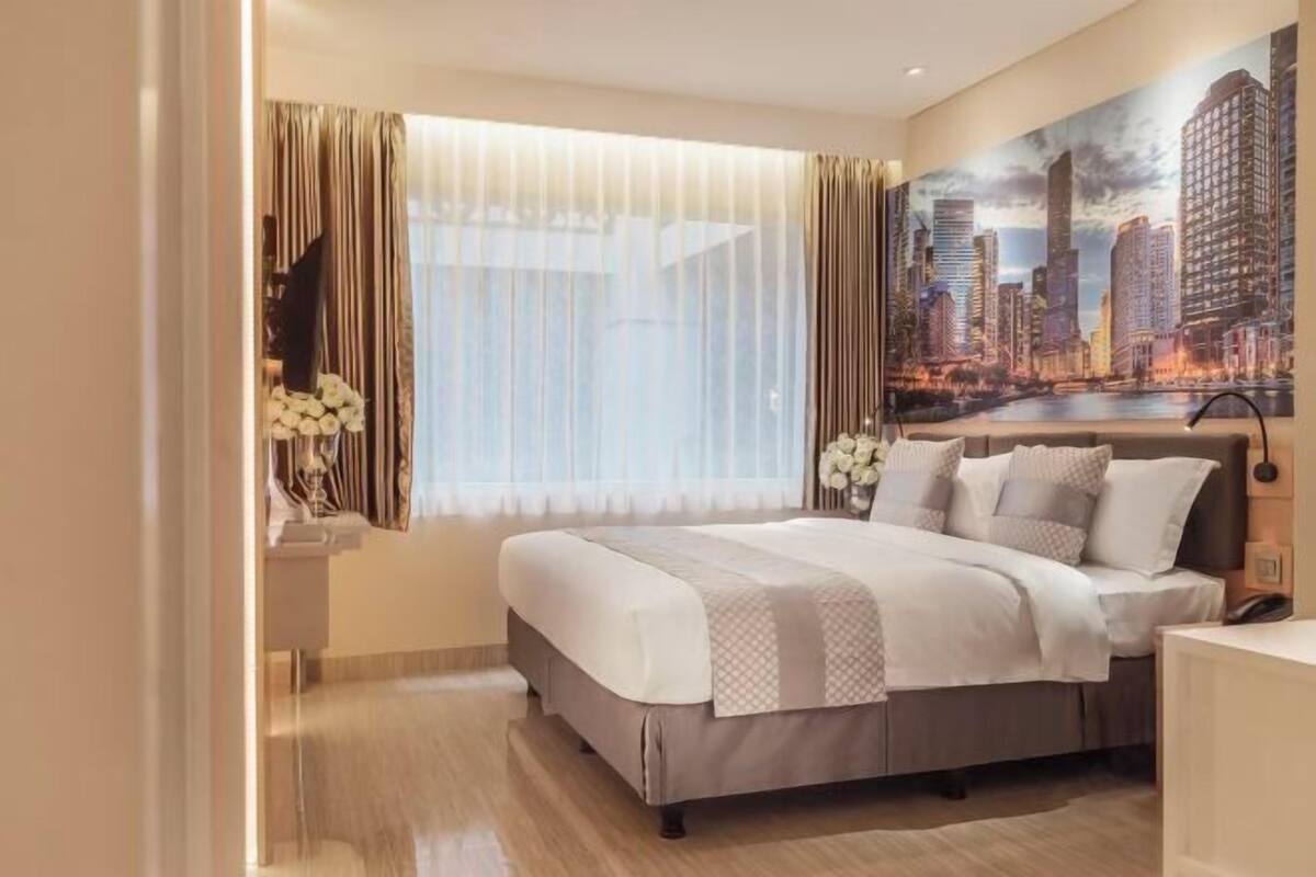 A hotel room with a large bed, a cityscape mural, a window with curtains, and a desk with a lamp.