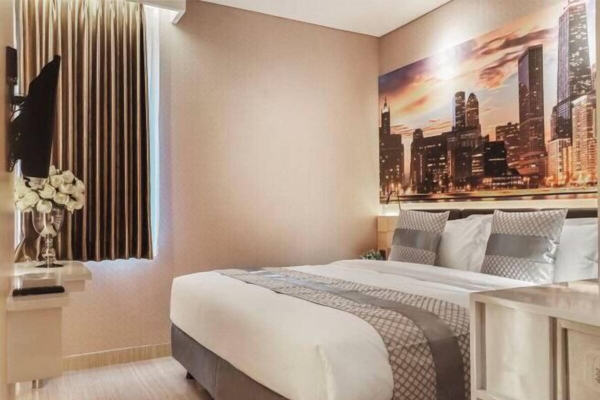 A hotel room with a bed, a TV, a window with curtains, and a cityscape painting on the wall.