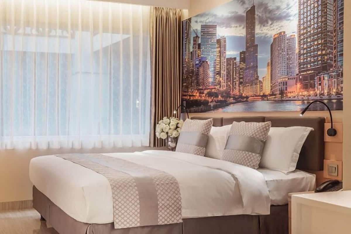 A hotel room with a large bed, a cityscape painting on the wall, and a view of the city through sheer curtains.