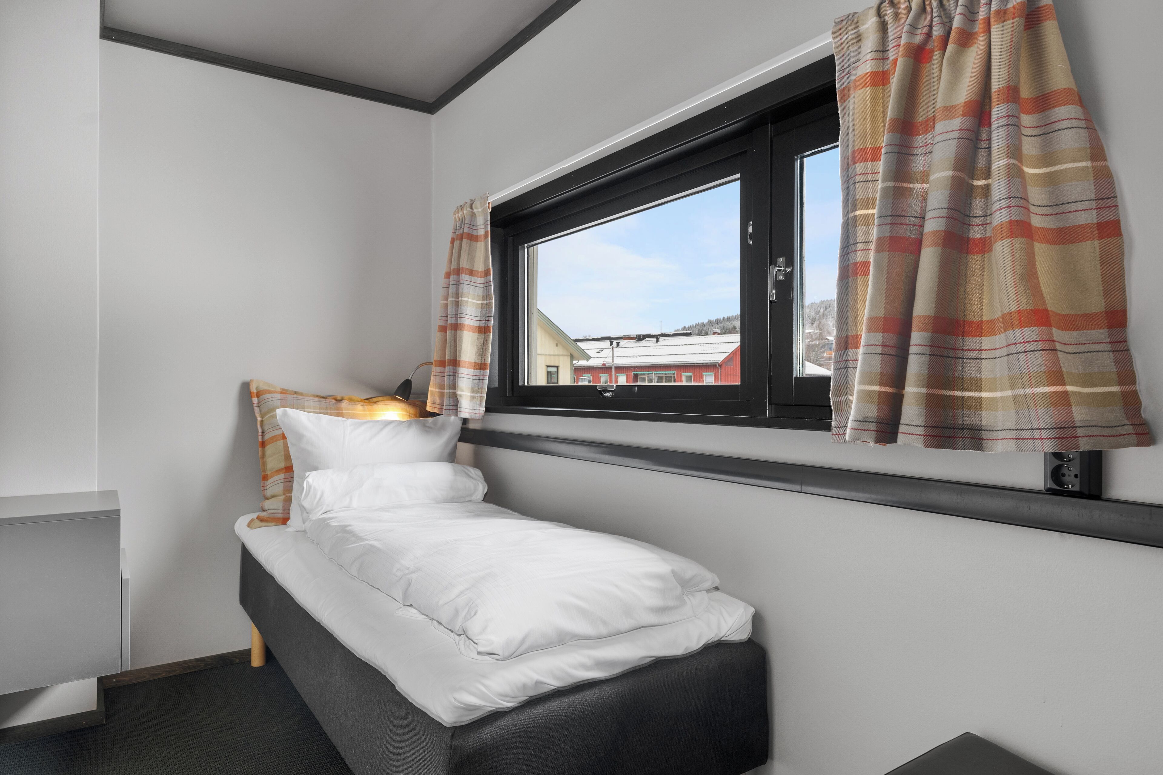 Une petite chambre avec un lit simple, une fenêtre et des rideaux, une table de chevet et une tête de lit.