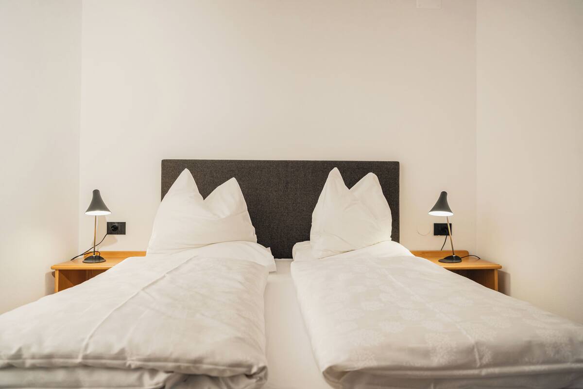 Uma cama de casal com um painel de cabeceira escuro, duas mesas de cabeceira com luminárias e uma parede lisa.