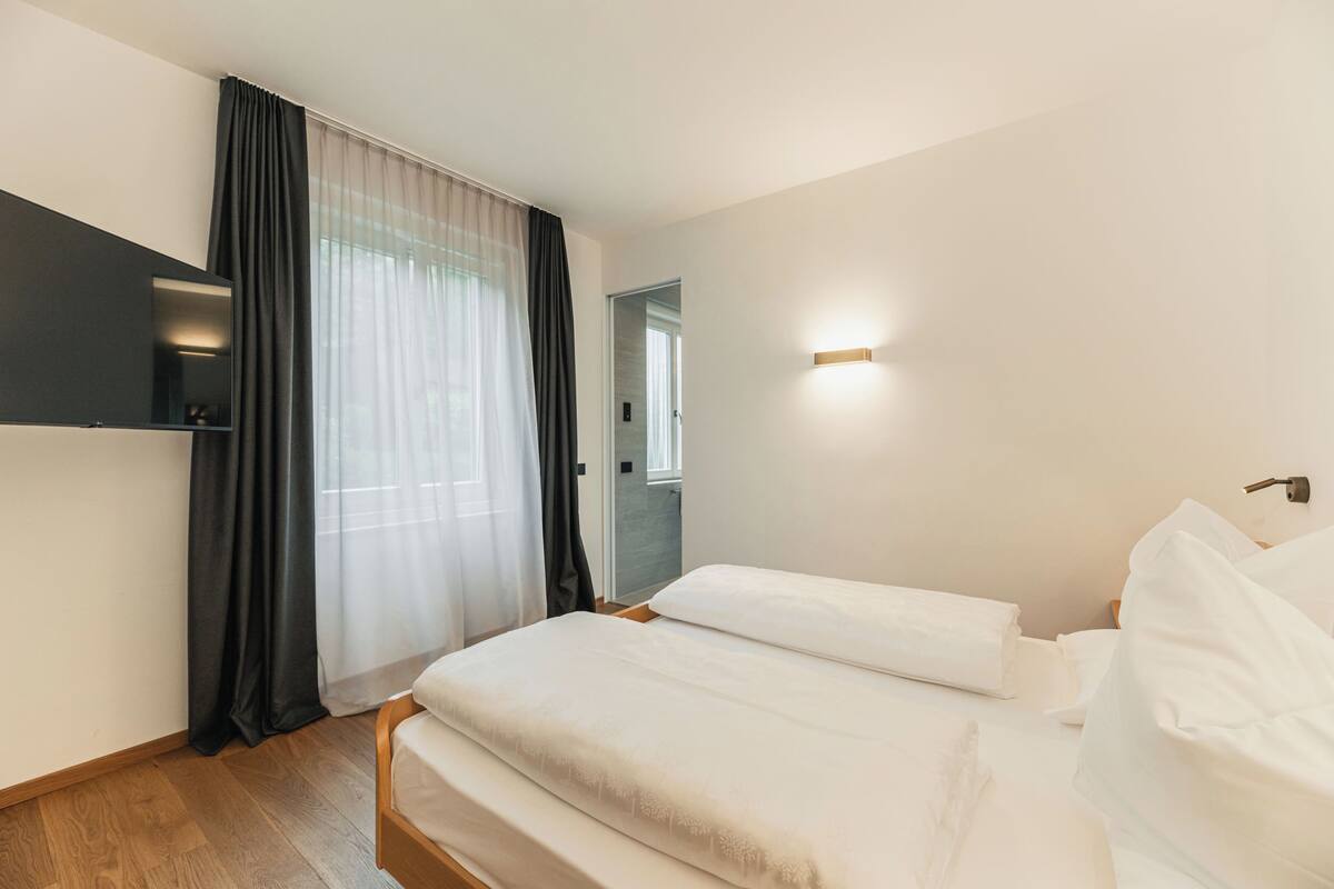 Quarto de hotel moderno com cama, televisão fixada na parede e janela com cortinas leves.