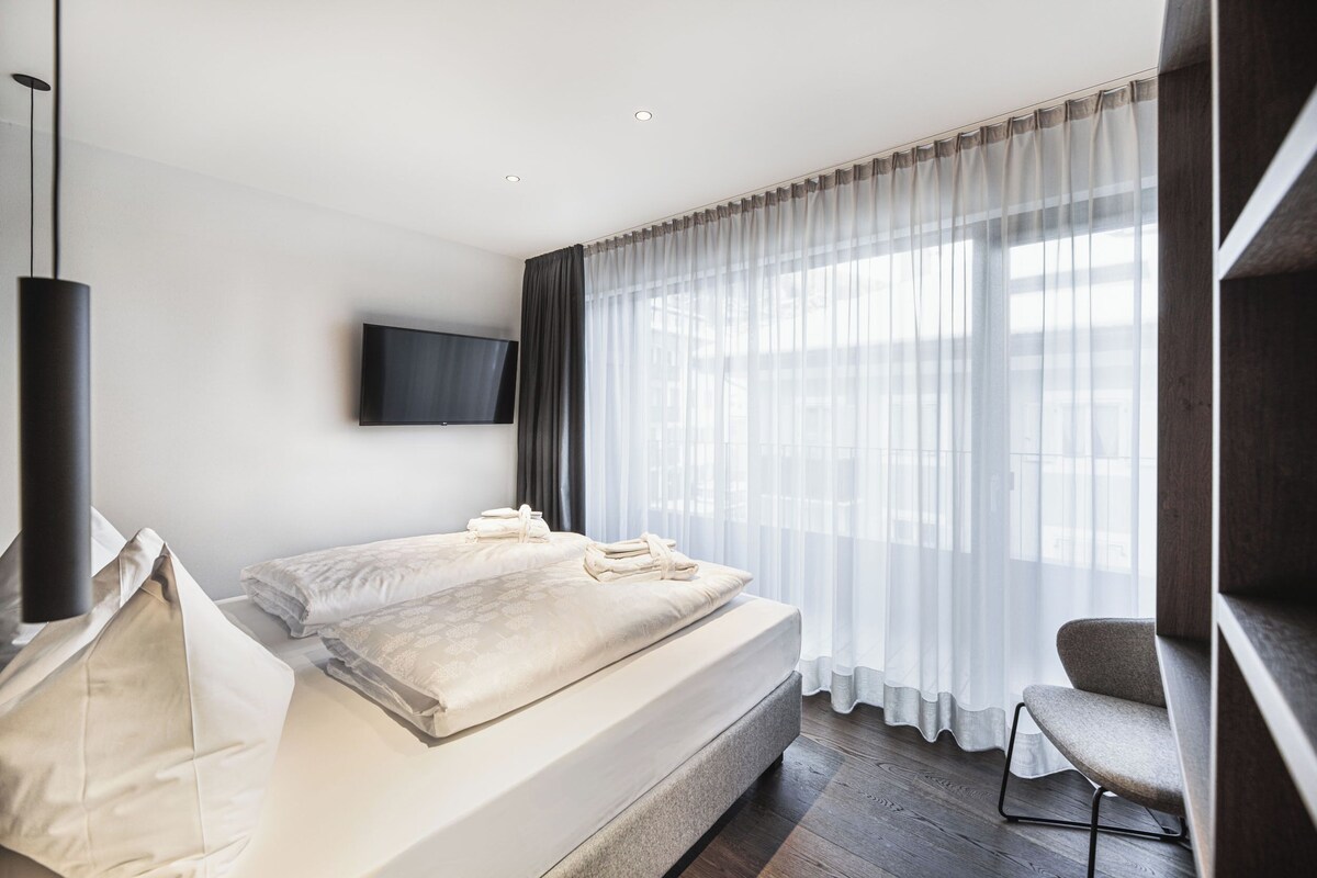 Une chambre moderne avec un grand lit, un téléviseur à écran plat fixé au mur et une grande fenêtre ornée de rideaux légers.