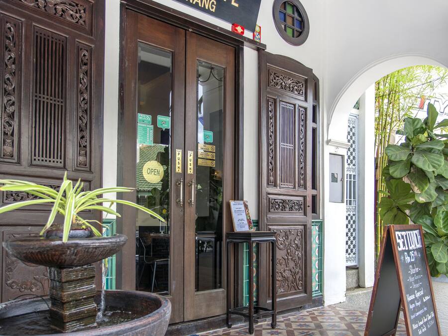 L'ingresso di un caffè con un cartello che indica "ARECA CAFE PENANG", una porta di legno decorativa, una piccola fontana e un pavimento con piastrelle decorate.