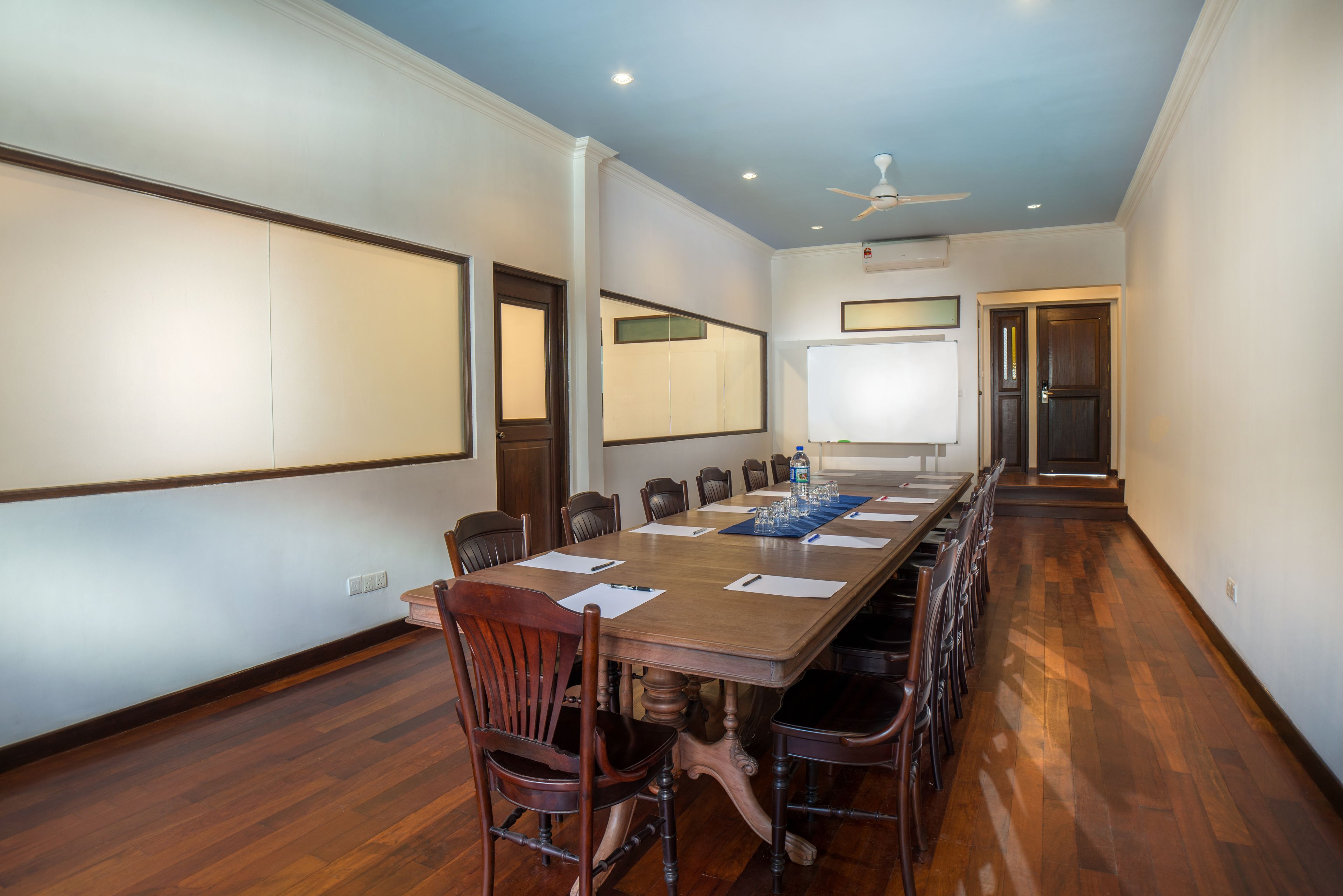 A conference room with a long wooden table, chairs, a whiteboard, and a ceiling fan.
