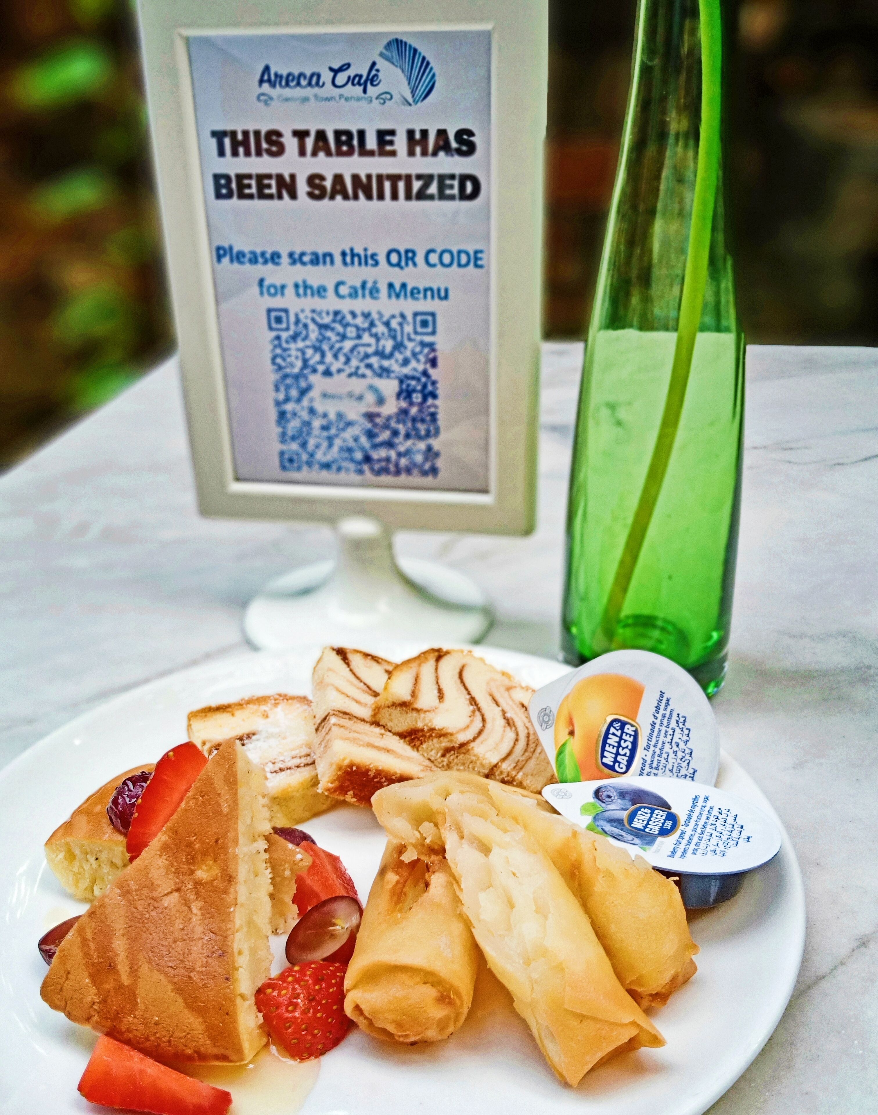 A table with a QR code sign for sanitization, a plate with a sandwich, fruit, and a drink.