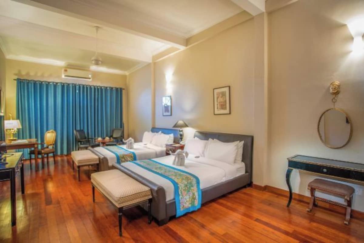 A hotel room with two beds, a desk, a chair, a bench, and a desk with a lamp.