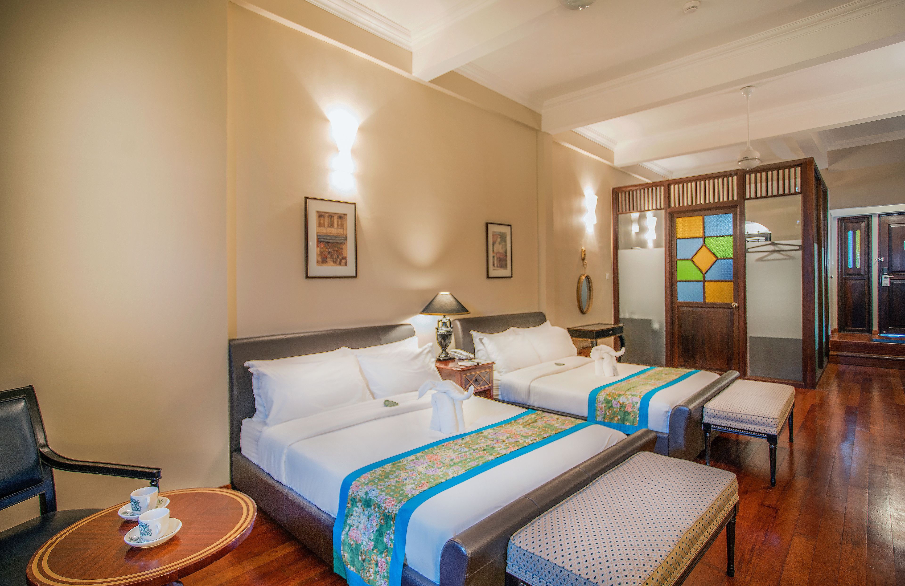 A hotel room with two beds, a desk with a chair, a small table with a cup and saucer, and a mirror with a colorful geometric pattern.