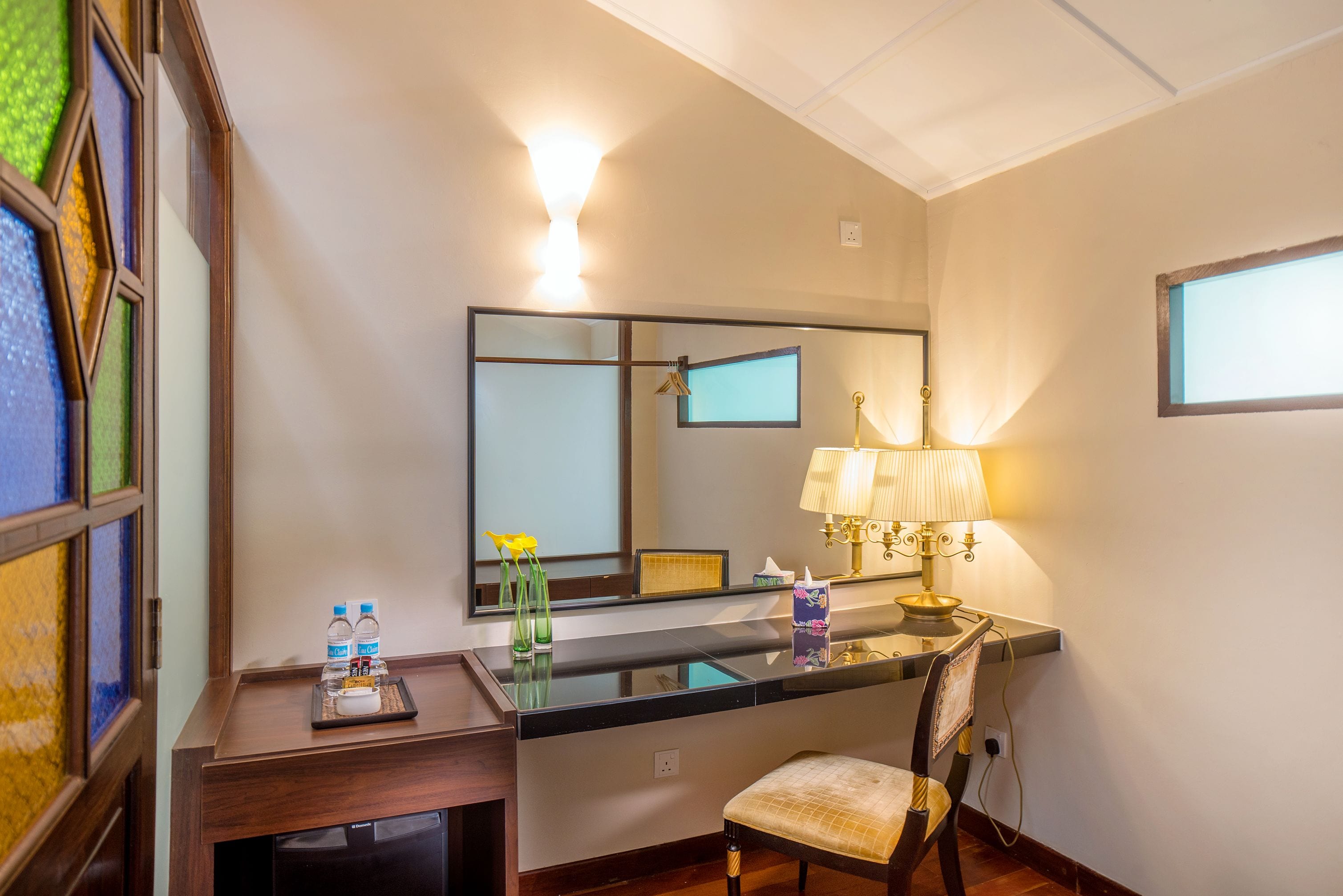 A hotel room with a desk, chair, and a large mirror. There is a lamp on the desk and a small table with a tray of toiletries.
