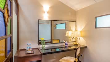 A hotel room with a desk, chair, and a large mirror. There is a lamp on the desk and a small table with a tray of toiletries.