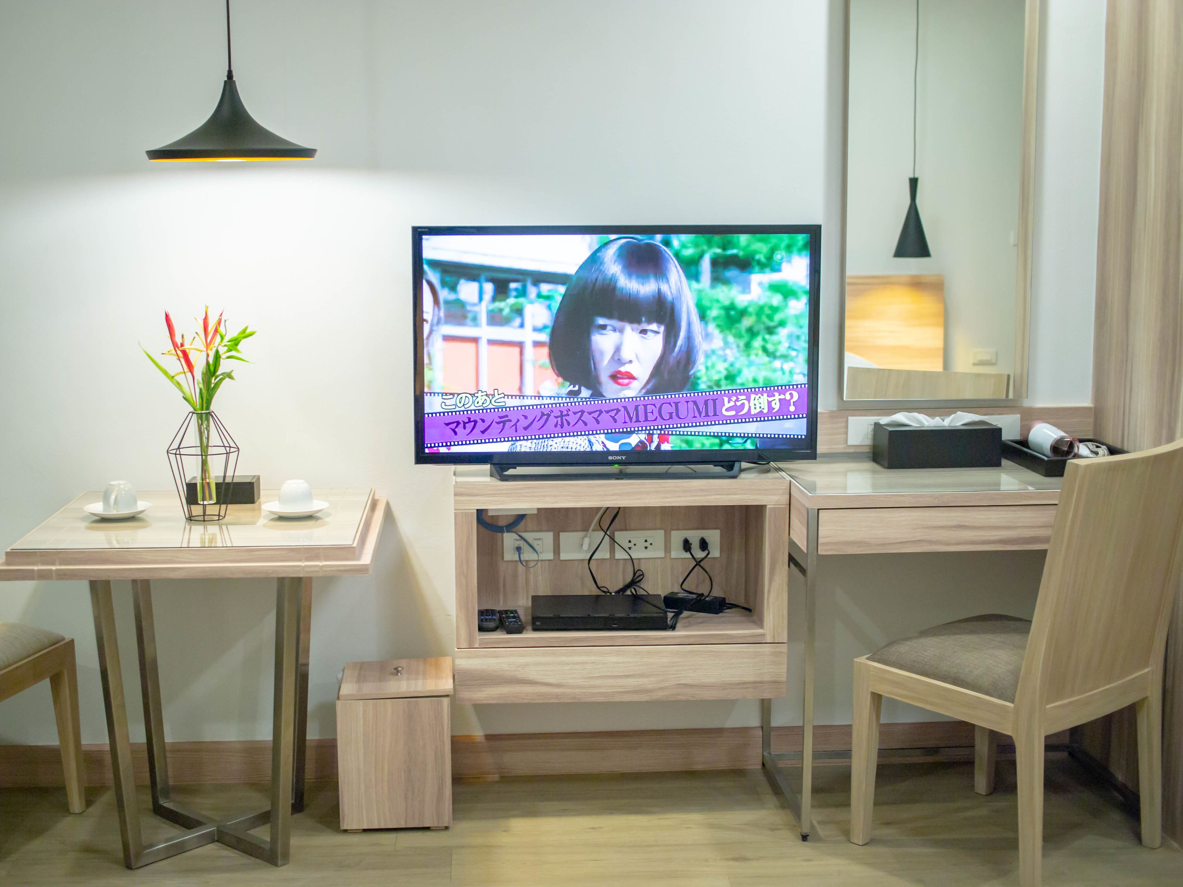 A modern hotel room with a flat-screen TV on a wooden stand, a small table with a vase of flowers, a chair, and a desk with a lamp.