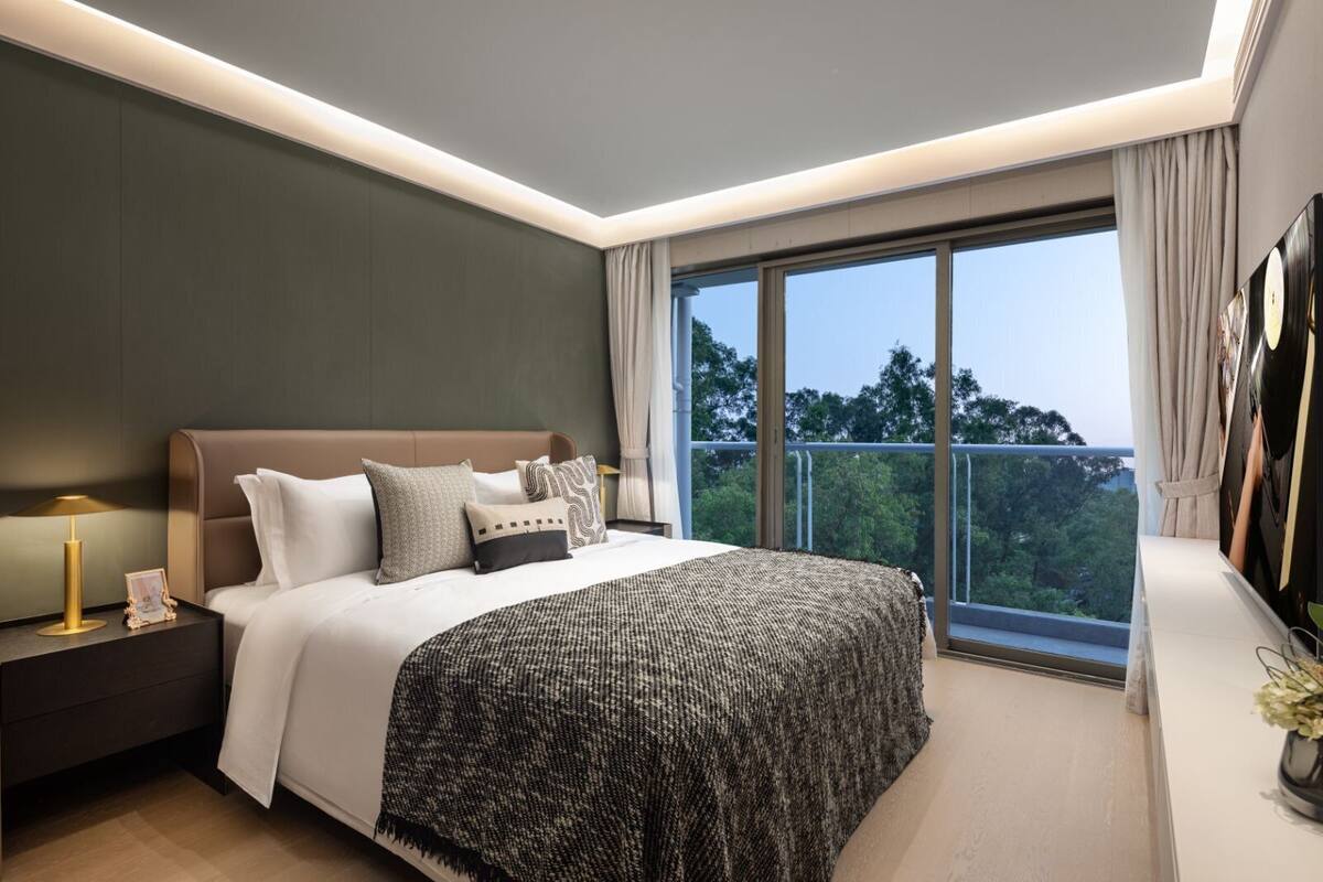 Ein modernes Schlafzimmer mit einem großen Bett, einem Nachttisch, einer Lampe und Blick ins Freie durch ein großes Fenster.