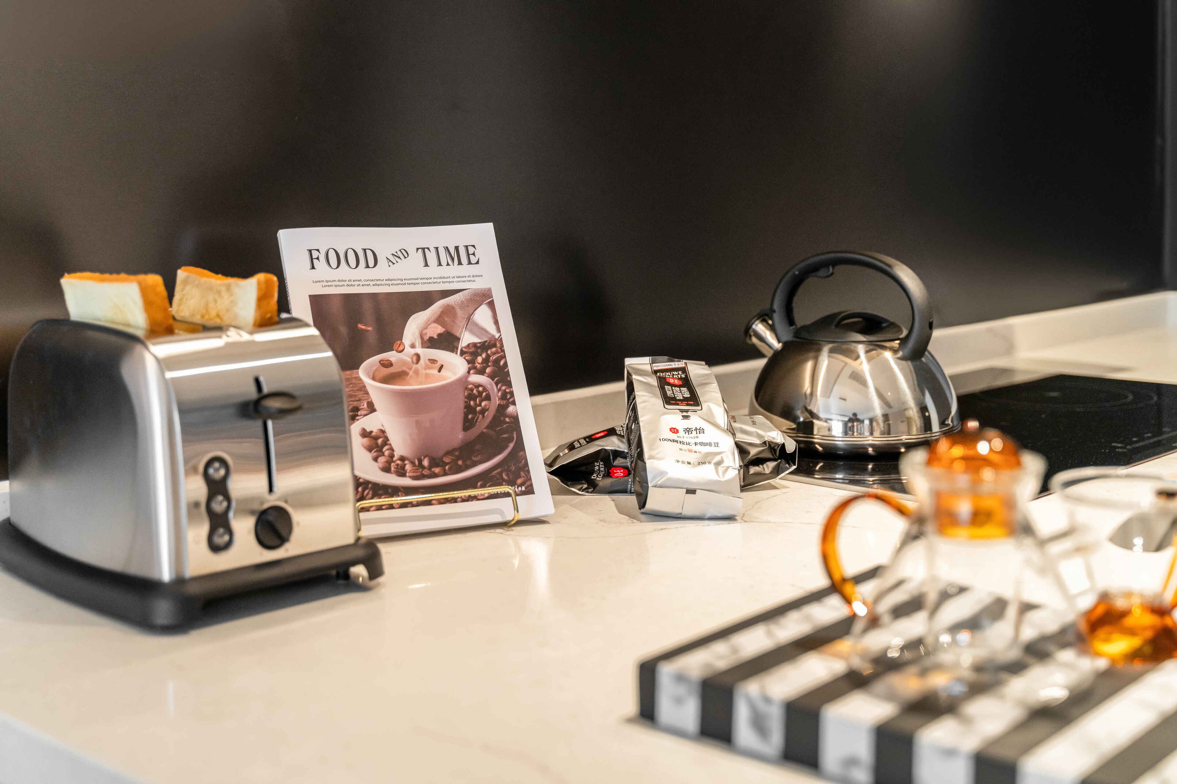 A kitchen counter with a toaster, a kettle, a teapot, a coffee pod, and a magazine titled "FOOD AND TIME".