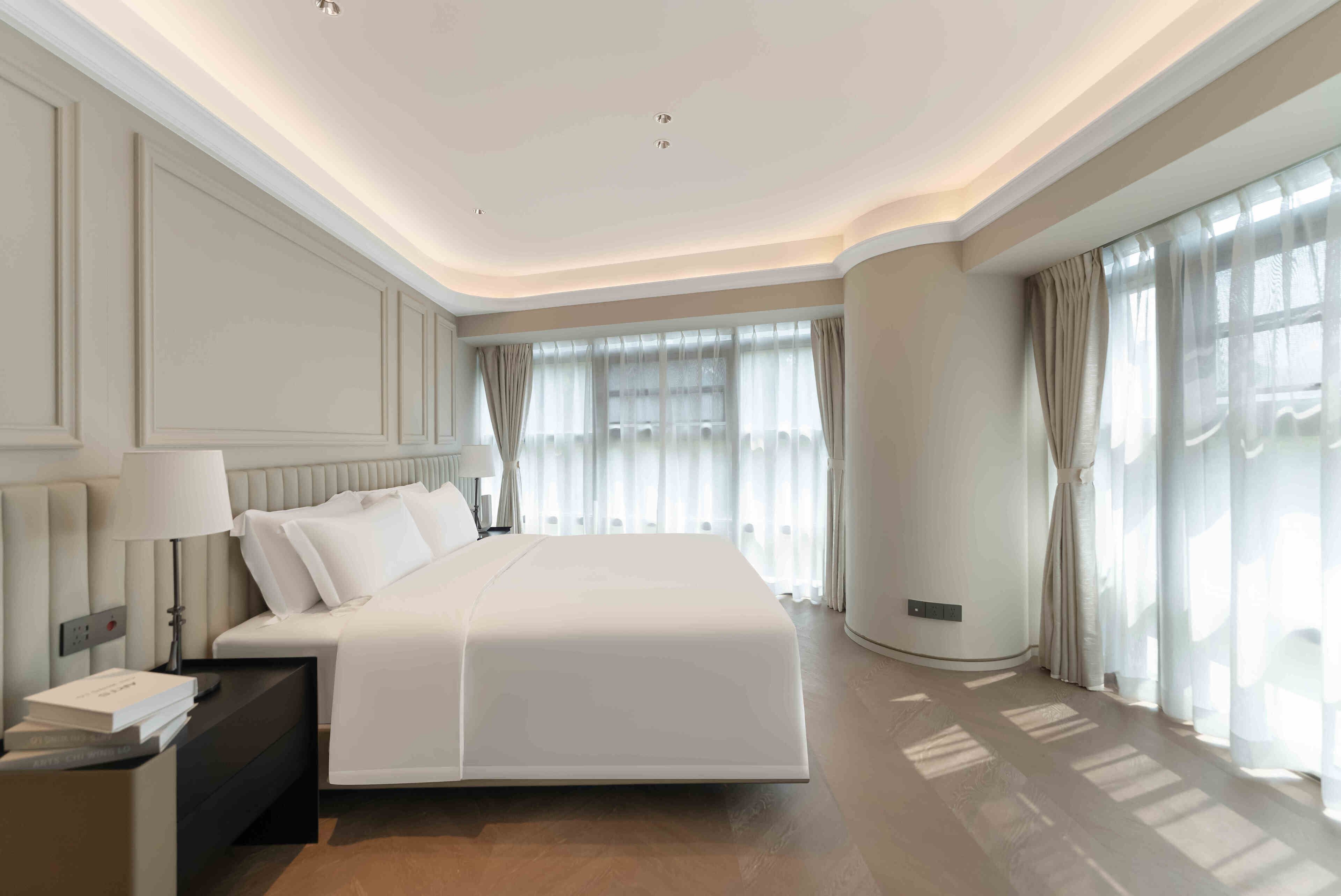 Una habitación de hotel moderna con una cama grande, una mesita de noche, una lámpara y vistas al exterior a través de amplias ventanas.