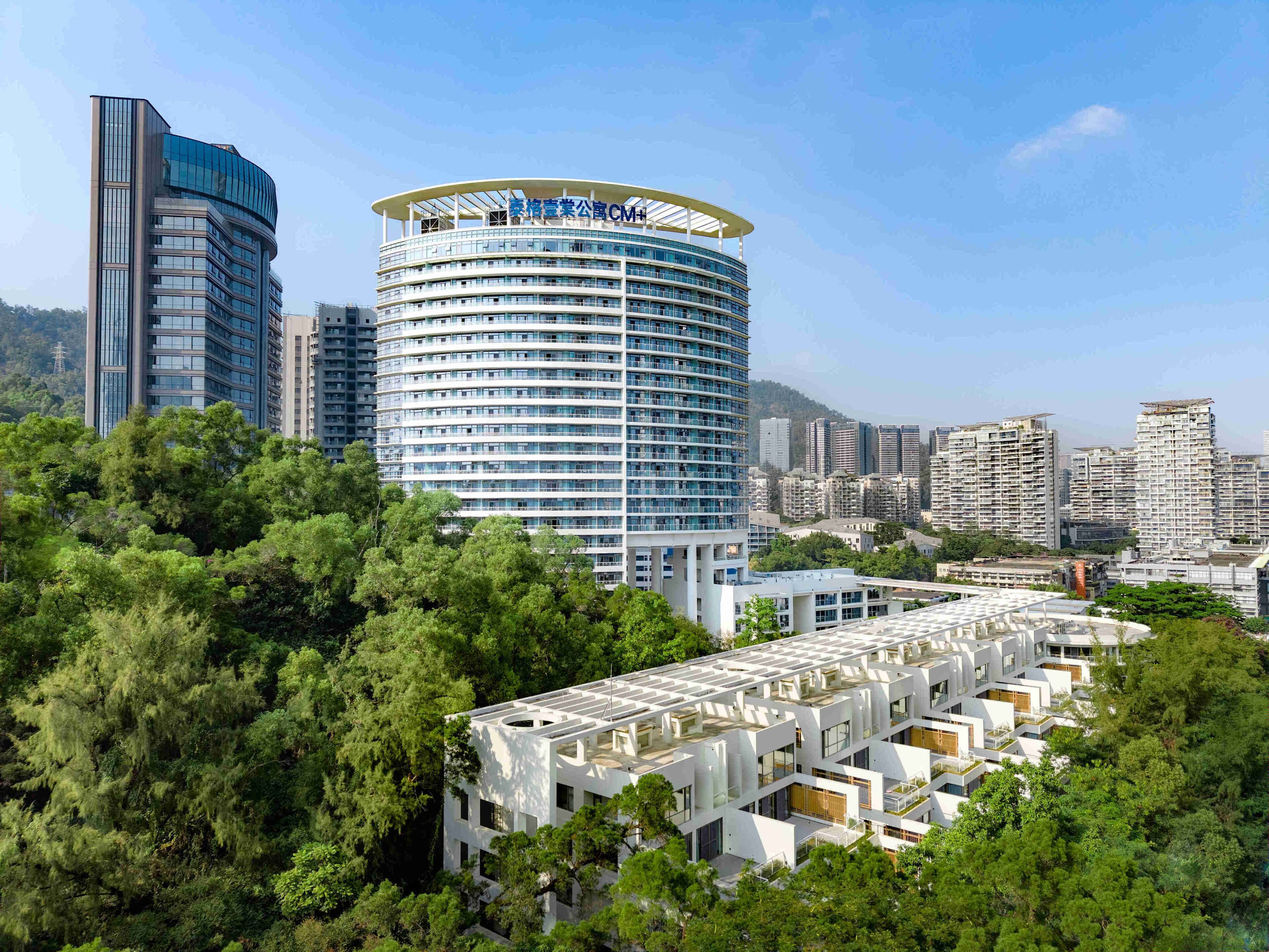A modern hotel with a circular design, surrounded by lush greenery and other high-rise buildings in the background.