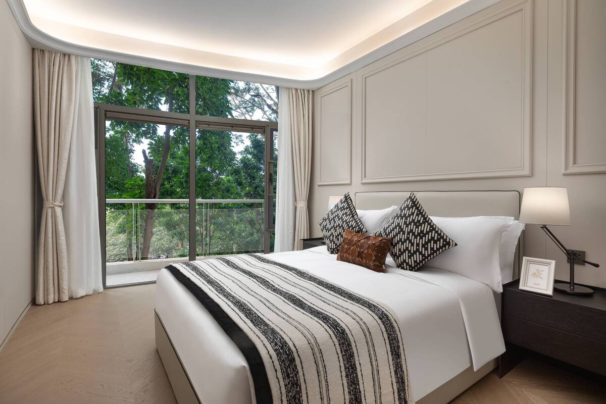 Ein modernes Hotelzimmer mit einem großen Bett, einem Nachttisch mit Lampe und Blick auf Grün durch ein großes Fenster.