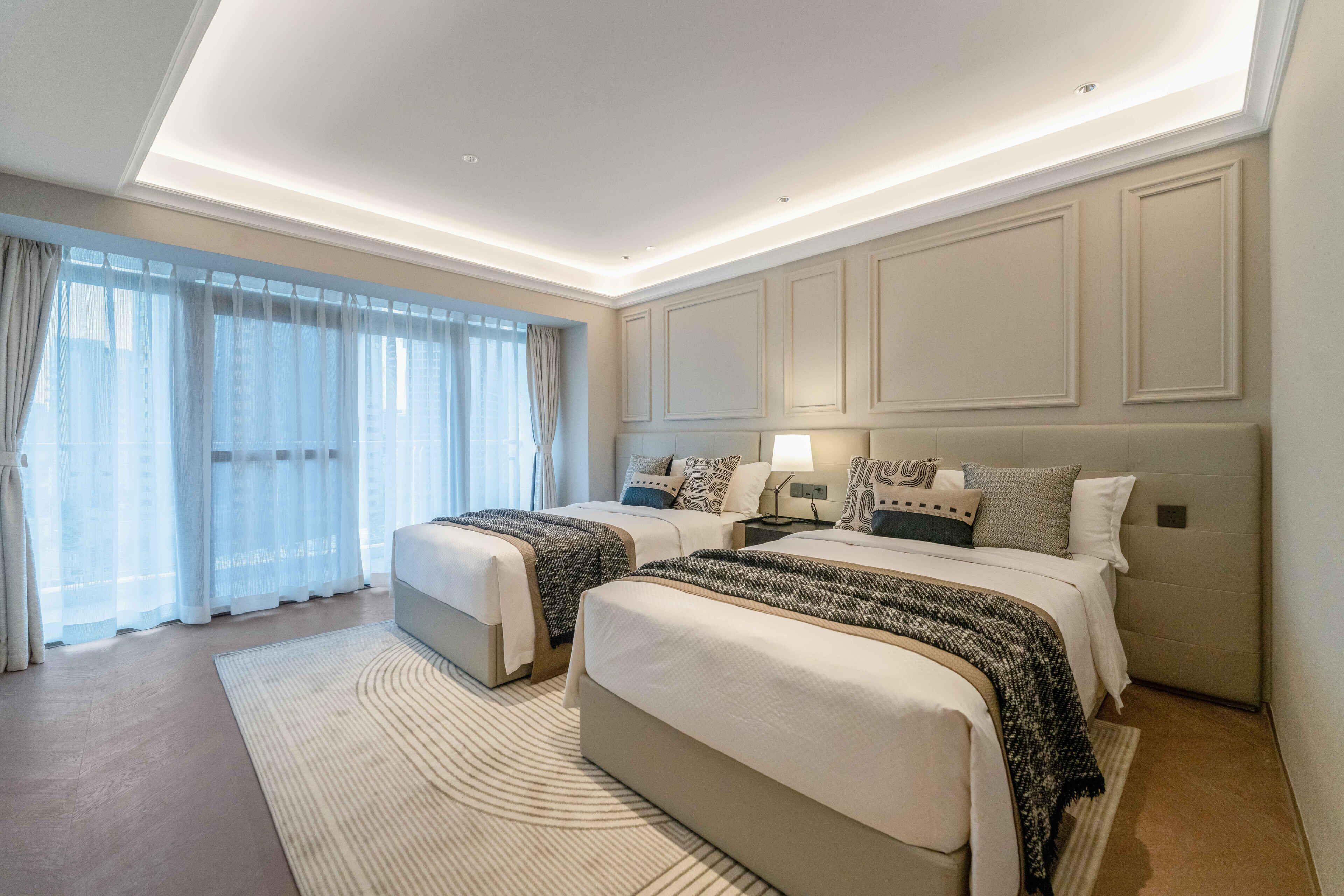 Una habitación de hotel moderna con dos camas, un ventanal con cortinas translúcidas e iluminación empotrada en el techo.