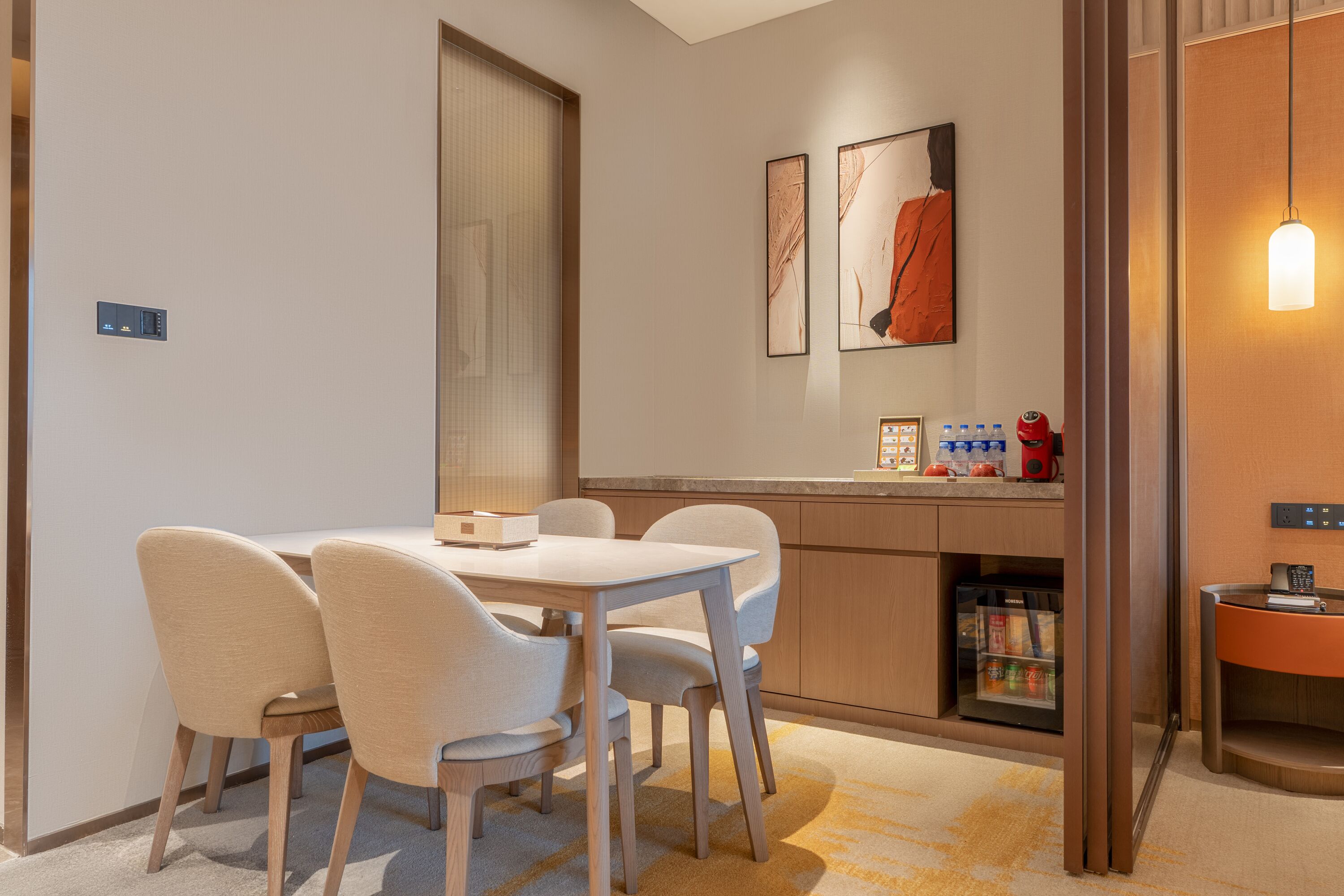 Uma área de refeições moderna com uma mesa e cadeiras, bancada de cozinha com eletrodomésticos e um pequeno espaço de estar com uma luminária.