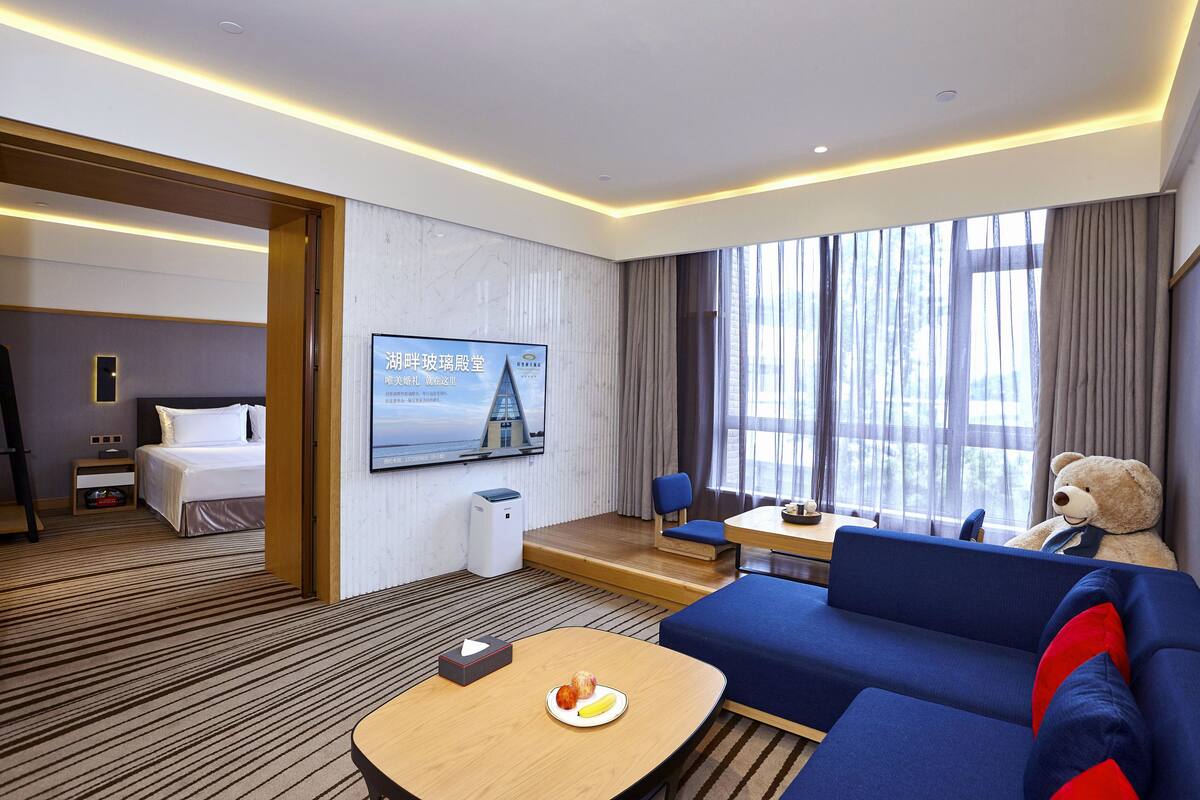 A hotel room with a bed, a TV, a sofa, a teddy bear, and a dining area with a plate of food.