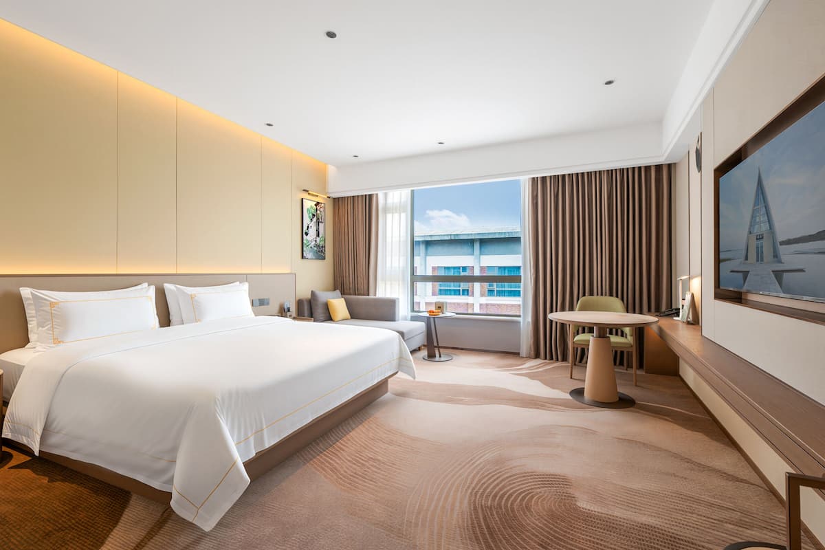 Quarto de hotel com uma cama grande, uma escrivaninha, uma cadeira, uma televisão e vista para um edifício pela janela.