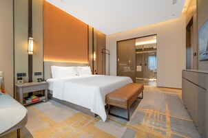 A modern hotel room with a large bed, bedside tables, a bench, a wardrobe, and a bathroom visible through a glass door.